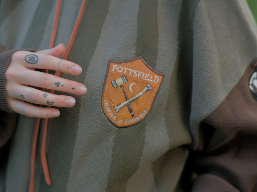 Cozy, cotton, knit hoodie featuring a striped body in shades of green, brown skeleton embroidered sleeves, and an orange pumpkin hood. The front features an embroidered harvest badge, and the back reads â€œyouâ€™ll join us somedayâ€.