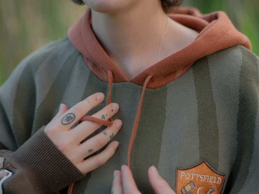 Cozy, cotton, knit hoodie featuring a striped body in shades of green, brown skeleton embroidered sleeves, and an orange pumpkin hood. The front features an embroidered harvest badge, and the back reads â€œyouâ€™ll join us somedayâ€.