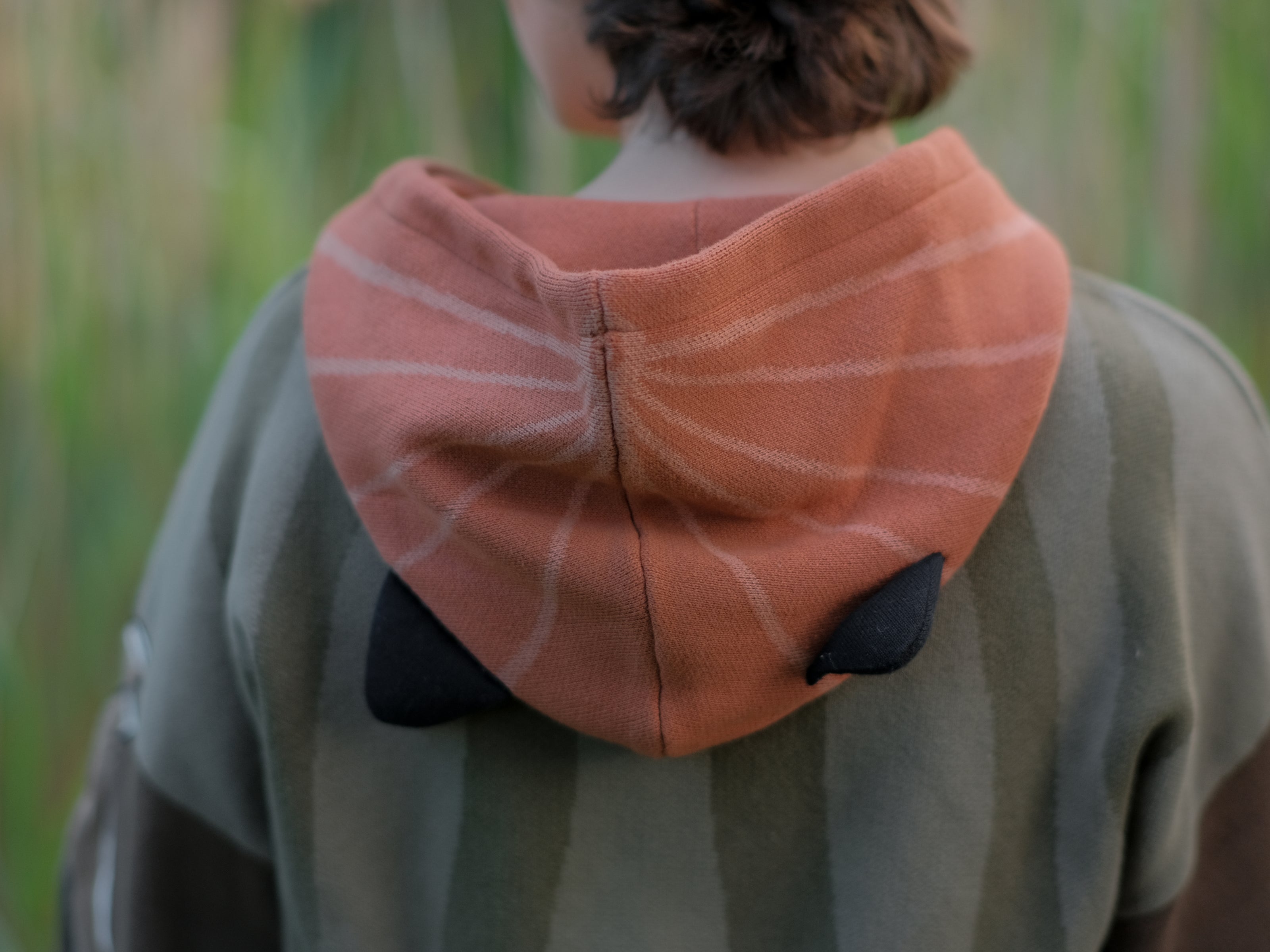 Cozy, cotton, knit hoodie featuring a striped body in shades of green, brown skeleton embroidered sleeves, and an orange pumpkin hood. The front features an embroidered harvest badge, and the back reads â€œyouâ€™ll join us somedayâ€.