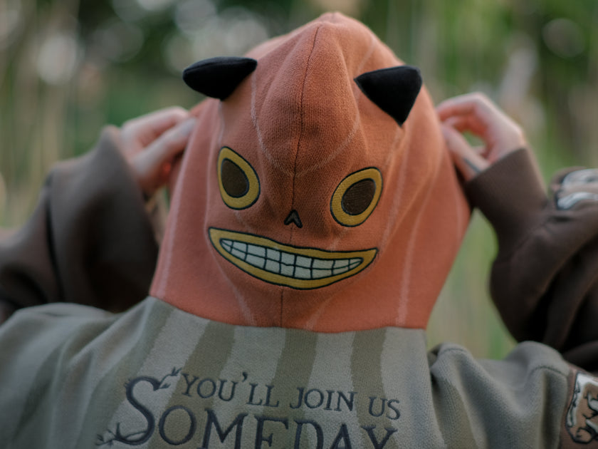 Cozy, cotton, knit hoodie featuring a striped body in shades of green, brown skeleton embroidered sleeves, and an orange pumpkin hood. The front features an embroidered harvest badge, and the back reads â€œyouâ€™ll join us somedayâ€.