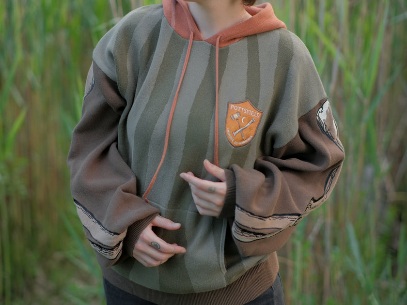 Cozy, cotton, knit hoodie featuring a striped body in shades of green, brown skeleton embroidered sleeves, and an orange pumpkin hood. The front features an embroidered harvest badge, and the back reads â€œyouâ€™ll join us somedayâ€.