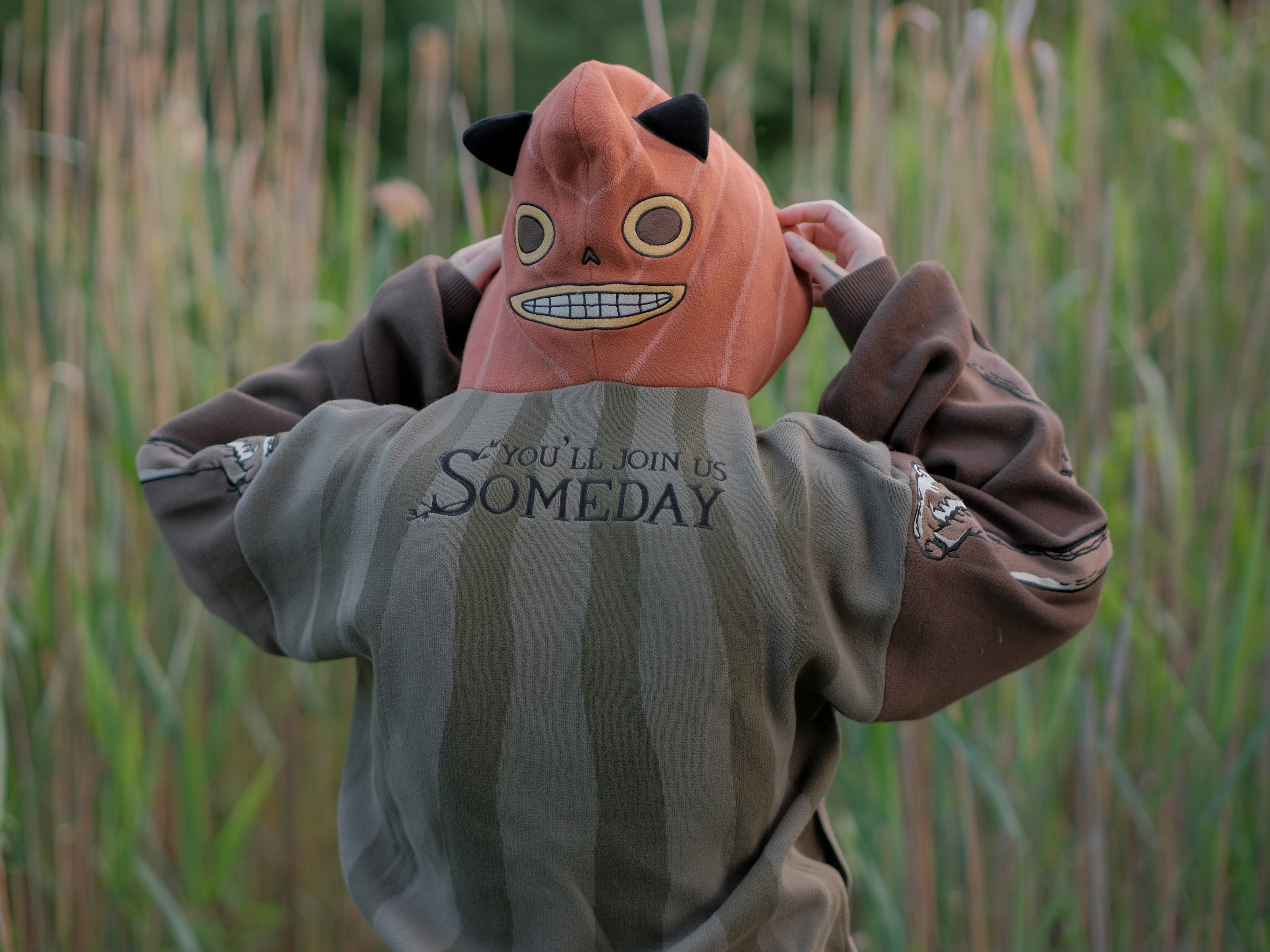 Cozy, cotton, knit hoodie featuring a striped body in shades of green, brown skeleton embroidered sleeves, and an orange pumpkin hood. The front features an embroidered harvest badge, and the back reads â€œyouâ€™ll join us somedayâ€.