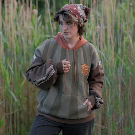 Cozy, cotton, knit hoodie featuring a striped body in shades of green, brown skeleton embroidered sleeves, and an orange pumpkin hood. The front features an embroidered harvest badge, and the back reads “you’ll join us someday”.