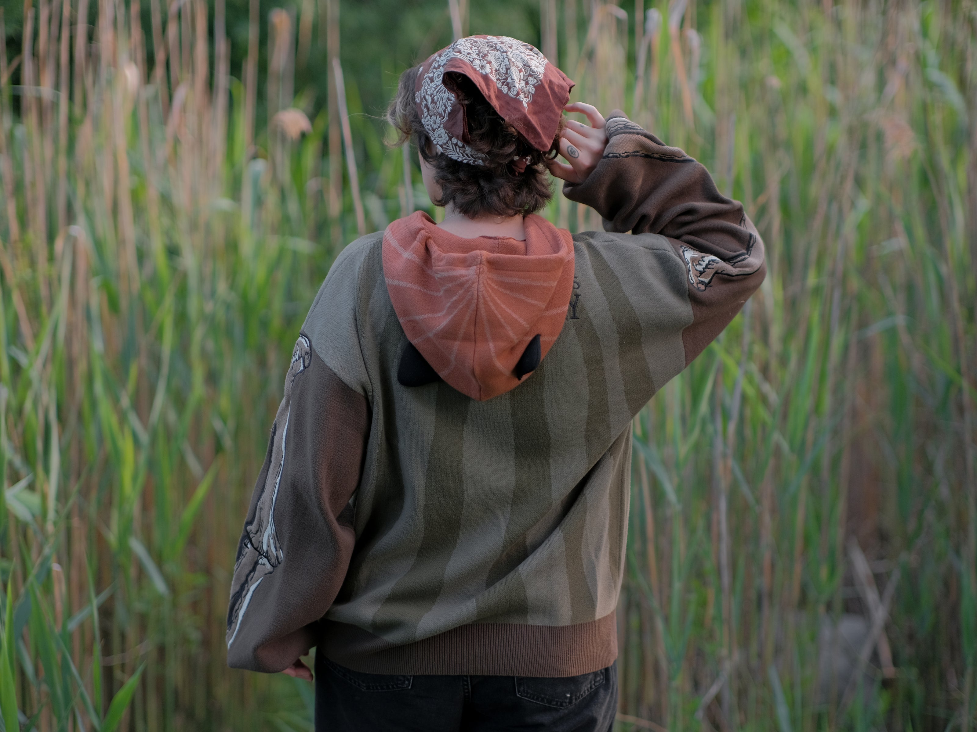 Cozy, cotton, knit hoodie featuring a striped body in shades of green, brown skeleton embroidered sleeves, and an orange pumpkin hood. The front features an embroidered harvest badge, and the back reads â€œyouâ€™ll join us somedayâ€.