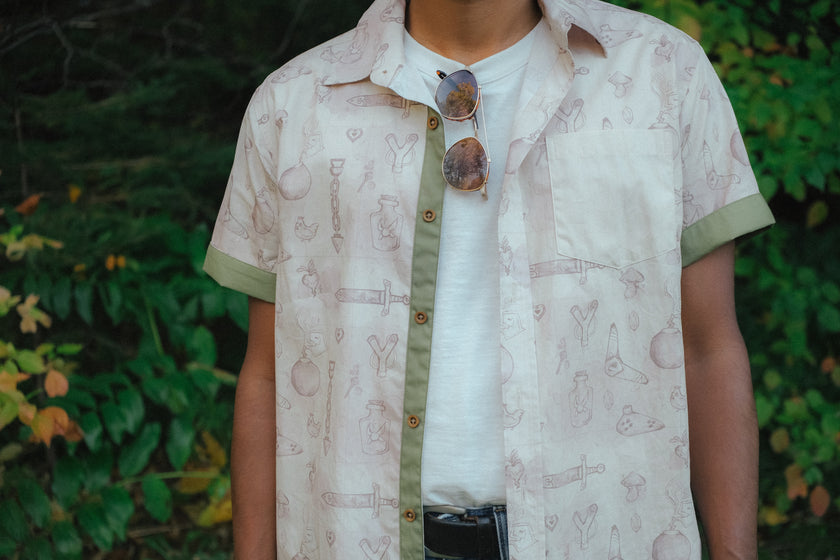 A person wearing a short-sleeve button-up shirt with a light beige and white abstract print, olive green trim, and brown buttons, layered over a white t-shirt with sunglasses hanging from the collar, set against a blurred green foliage background.