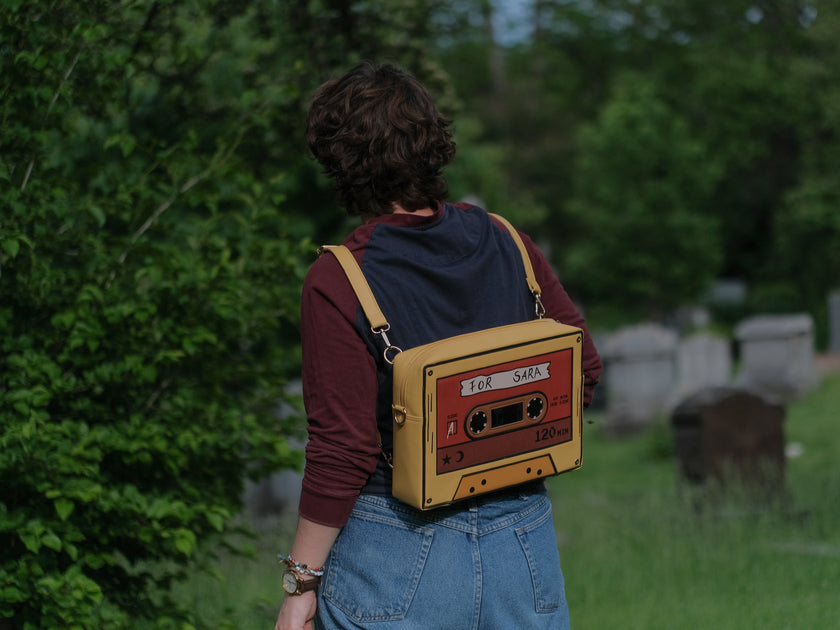 Orange and yellow backpack/satchel, designed to resemble a mix-tape, featuring the classic tape shape, and reads “For Sarah”. The bag includes 2 adjustable backpack straps and 1 adjustable cross-body strap, and features two inner pockets, a padded section, and pen straps. Inner fabric is custom-printed with a stain-resistant fabric.