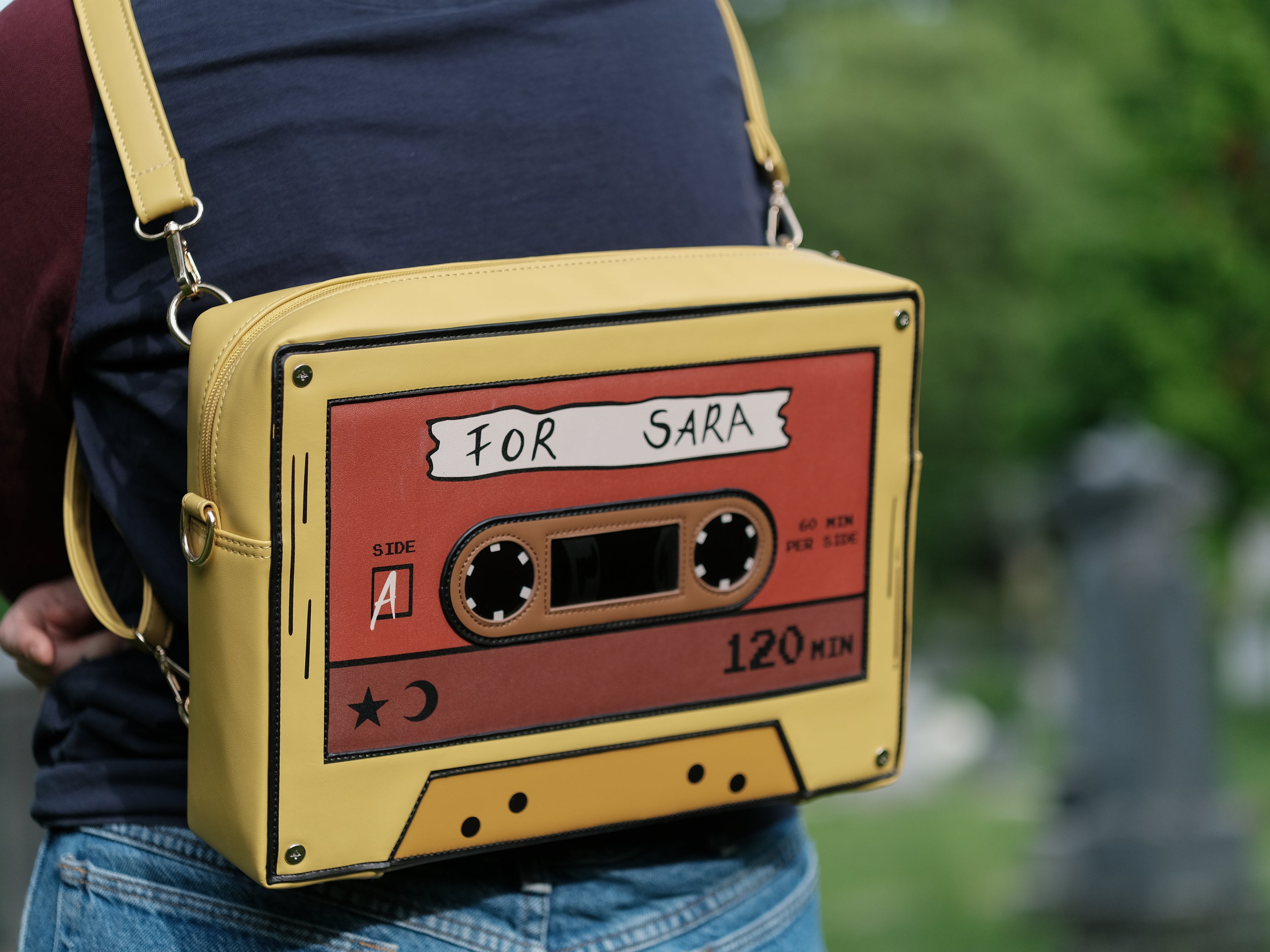 Orange and yellow backpack/satchel, designed to resemble a mix-tape, featuring the classic tape shape, and reads “For Sarah”. The bag includes 2 adjustable backpack straps and 1 adjustable cross-body strap, and features two inner pockets, a padded section, and pen straps. Inner fabric is custom-printed with a stain-resistant fabric.