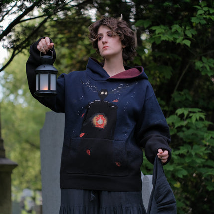 Cozy, cotton, knit hoodie in black and navy with red lining. The sweater features an intricately printed design of a beastly woodland creature, and is detailed with embroidered leaves, glowing eyes, melting snow, and lantern.