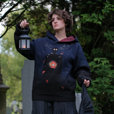 Cozy, cotton, knit hoodie in black and navy with red lining. The sweater features an intricately printed design of a beastly woodland creature, and is detailed with embroidered leaves, glowing eyes, melting snow, and lantern.