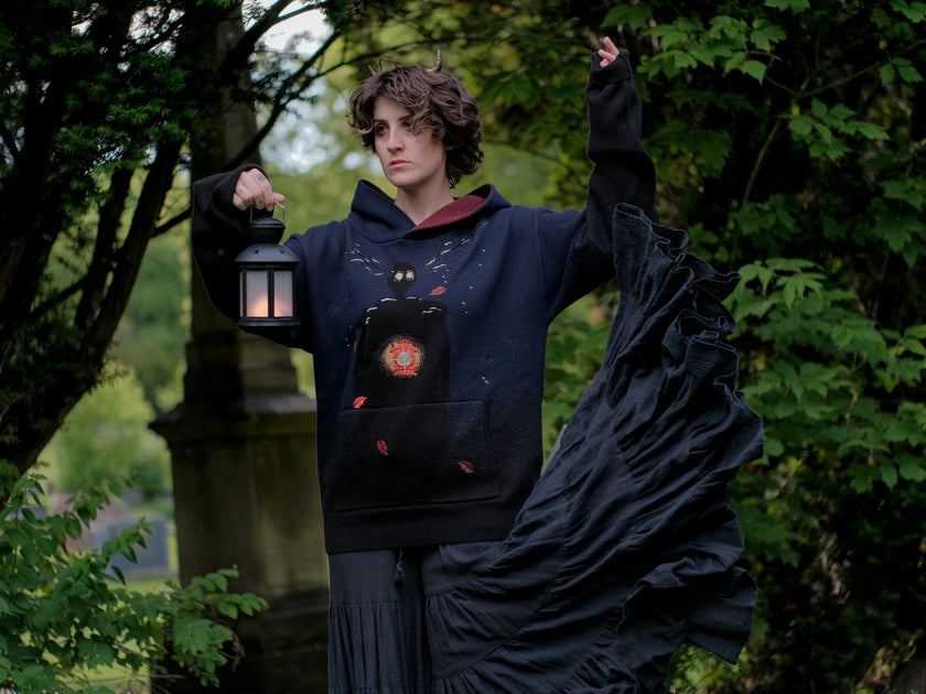 Cozy, cotton, knit hoodie in black and navy with red lining. The sweater features an intricately printed design of a beastly woodland creature, and is detailed with embroidered leaves, glowing eyes, melting snow, and lantern.
