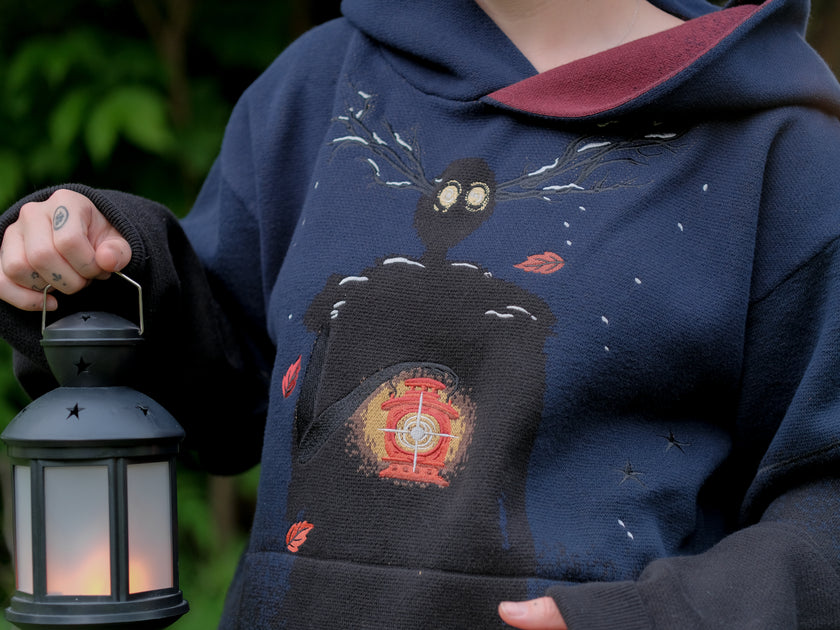 Cozy, cotton, knit hoodie in black and navy with red lining. The sweater features an intricately printed design of a beastly woodland creature, and is detailed with embroidered leaves, glowing eyes, melting snow, and lantern.