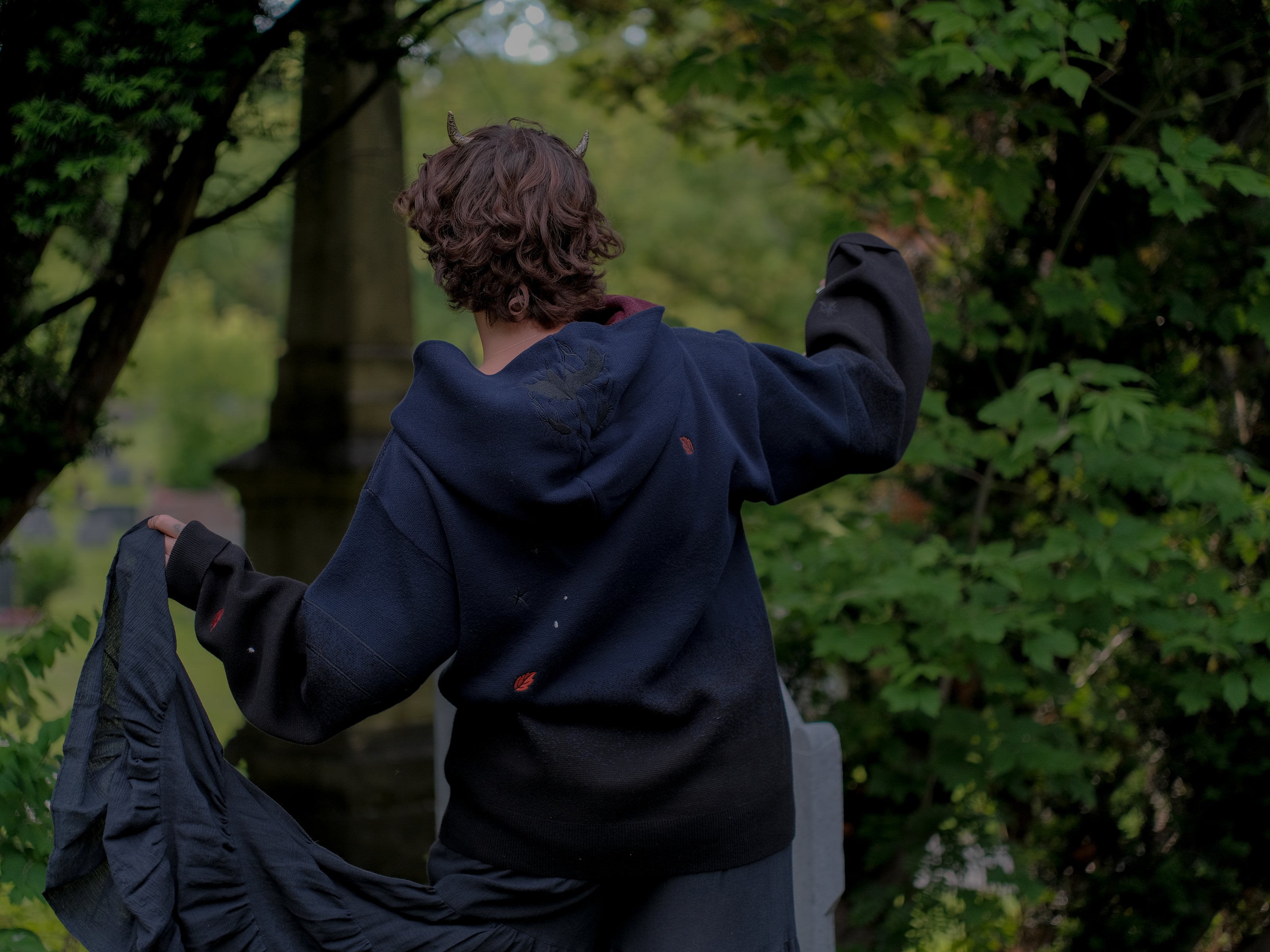 Cozy, cotton, knit hoodie in black and navy with red lining. The sweater features an intricately printed design of a beastly woodland creature, and is detailed with embroidered leaves, glowing eyes, melting snow, and lantern.