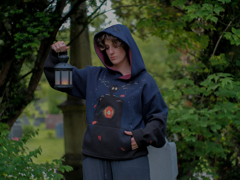 Cozy, cotton, knit hoodie in black and navy with red lining. The sweater features an intricately printed design of a beastly woodland creature, and is detailed with embroidered leaves, glowing eyes, melting snow, and lantern.