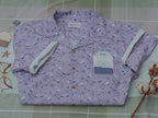 A muted purple, short-sleeve button up t-shirt printed with a whimsical pattern of fancy teacups, bergamot flowers, lavender sprigs, tea bags, spoons and sugar cubes. The shirt has a pyramid tea bag pocket, as well as a relaxed fit and rolled cuffs.