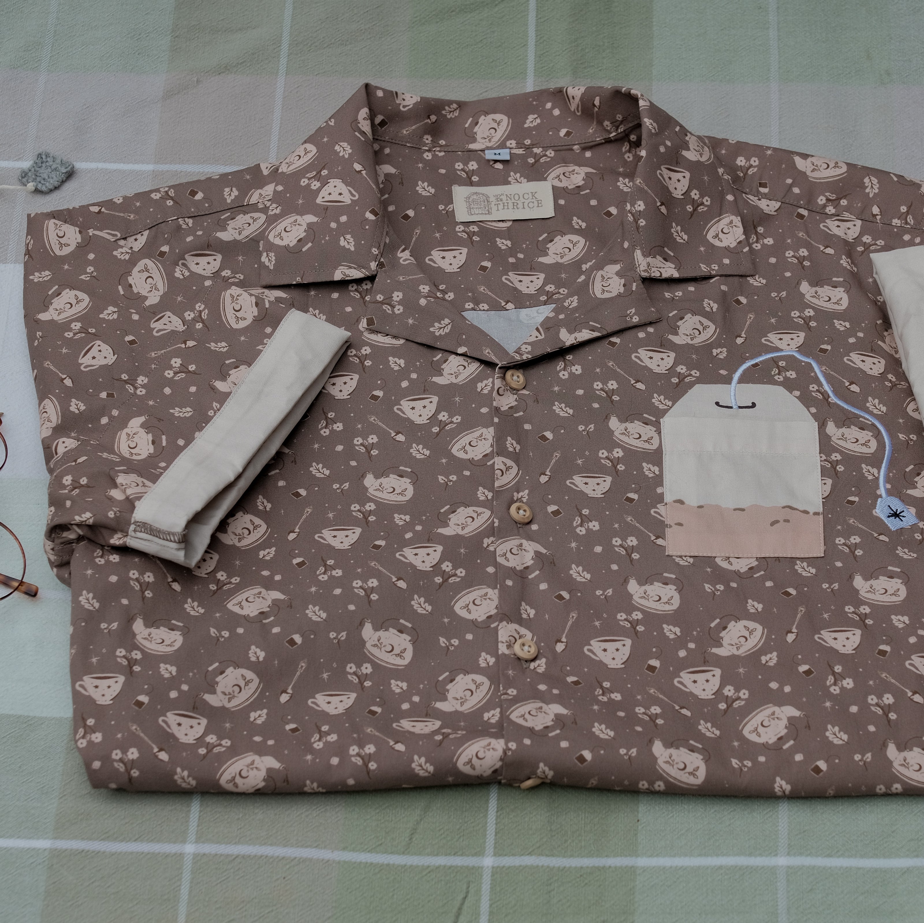 Warm, boba brown shade short-sleeve button up t-shirt printed with a whimsical pattern of the classic chubby teapot and matching cup, autumnal oak leaves, tea bag, spoon and sugar cubes. The shirt has a pyramid tea bag pocket, as well as a relaxed fit and rolled cuffs.