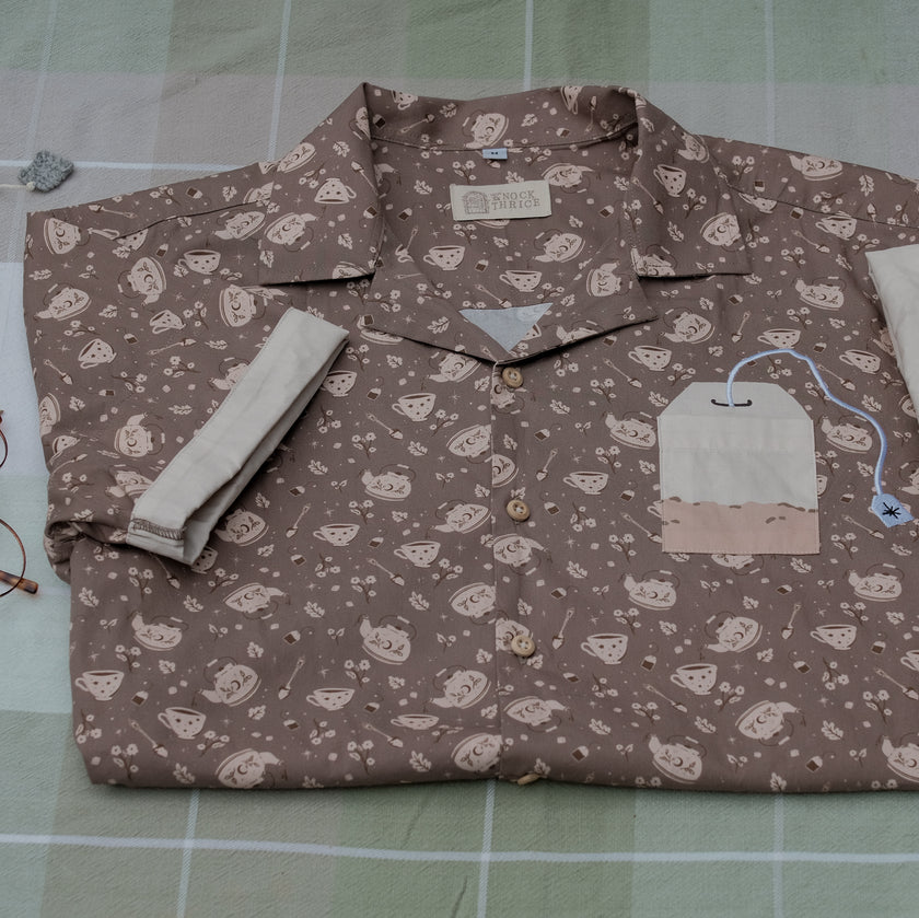 Warm, boba brown shade short-sleeve button up t-shirt printed with a whimsical pattern of the classic chubby teapot and matching cup, autumnal oak leaves, tea bag, spoon and sugar cubes. The shirt has a pyramid tea bag pocket, as well as a relaxed fit and rolled cuffs.