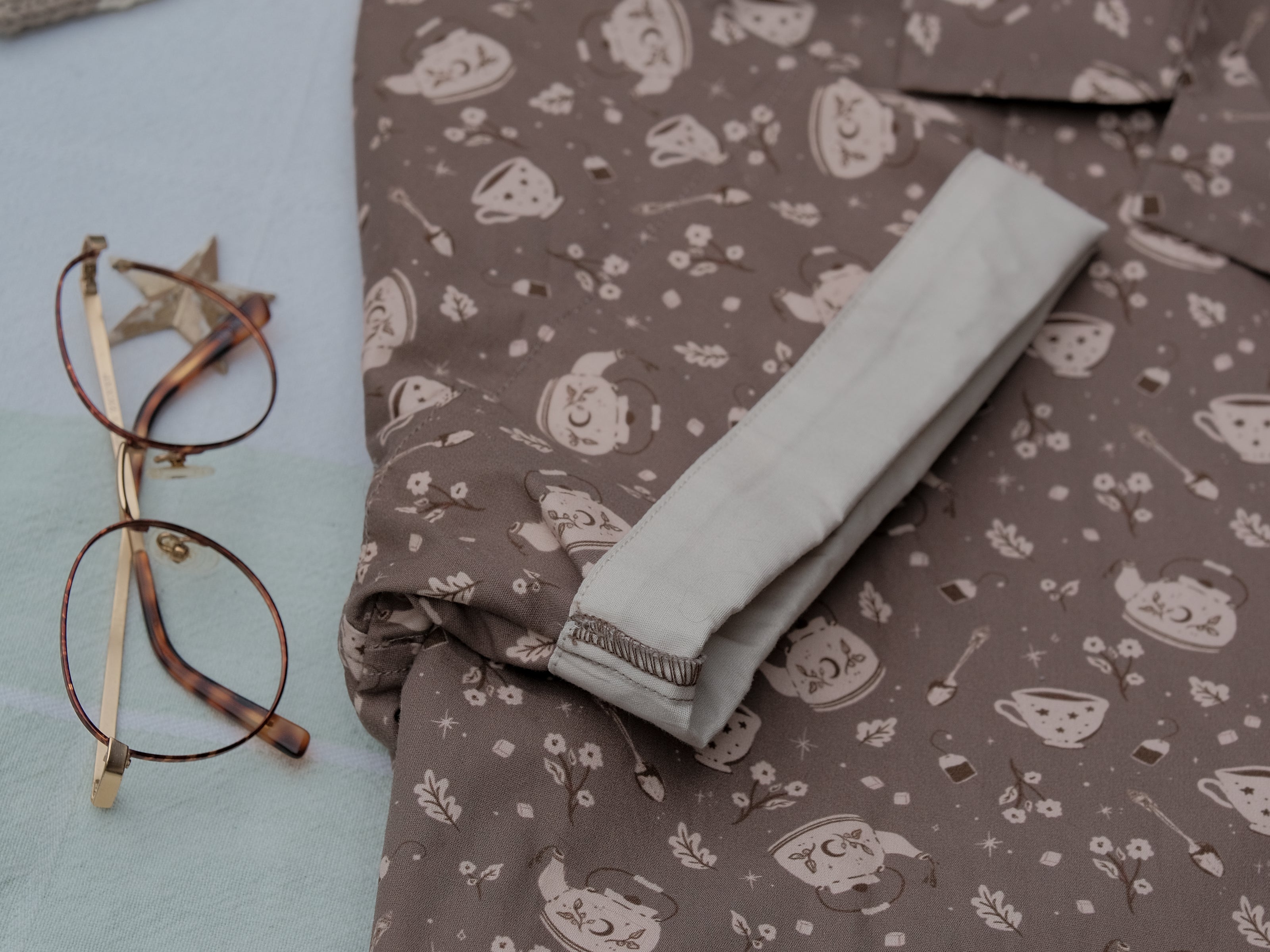 Warm, boba brown shade short-sleeve button up t-shirt printed with a whimsical pattern of the classic chubby teapot and matching cup, autumnal oak leaves, tea bag, spoon and sugar cubes. The shirt has a pyramid tea bag pocket, as well as a relaxed fit and rolled cuffs.