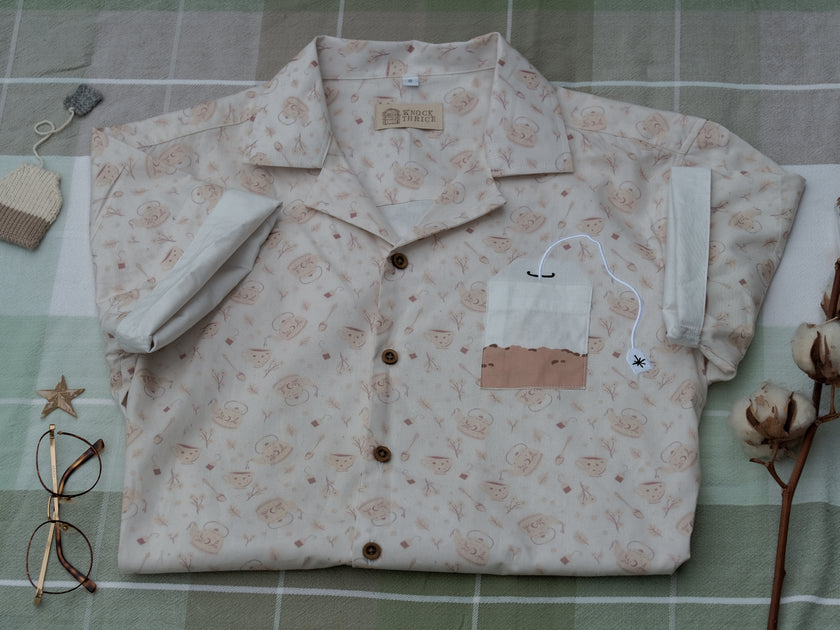 A warm ivory/cream colour short-sleeve button up t-shirt printed with a whimsical pattern of classic chubby teapots and matching cups, autumnal oak leaves, tea bags, spoons and sugar cubes. The shirt has a pyramid tea bag pocket, as well as a relaxed fit and rolled cuffs.