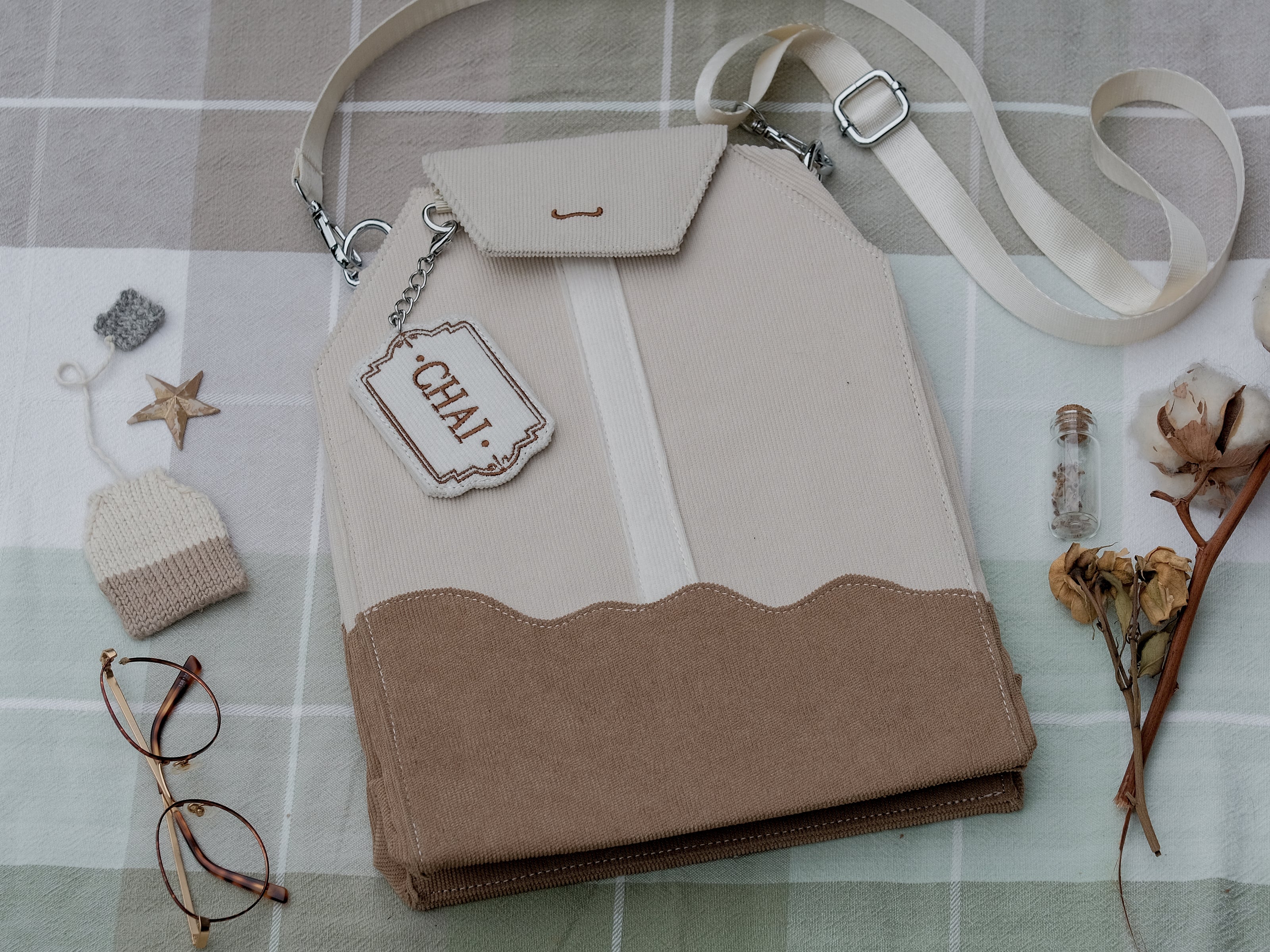 Ivory/cream and warm brown cotton corduroy purse, designed to resemble a chai tea bag, featuring the classic pyramid tea bag shape, adjustable matching strap, keychain rings, and a tag detail hanging from a strap. The bag features two inner pockets, one zipper and one padded, as well as 2 other unpadded pockets. Inner fabric is printed with a pattern of tea pots, bags, spoons, and cups, oak leaves, flowers, and stars on a soft brown background.