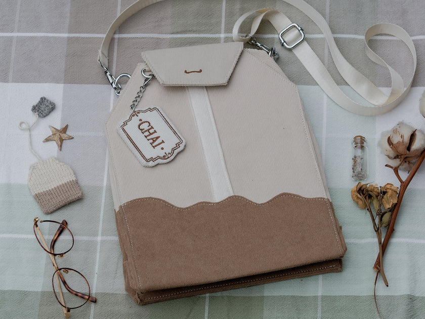 Ivory/cream and warm brown cotton corduroy purse, designed to resemble a chai tea bag, featuring the classic pyramid tea bag shape, adjustable matching strap, keychain rings, and a tag detail hanging from a strap. The bag features two inner pockets, one zipper and one padded, as well as 2 other unpadded pockets. Inner fabric is printed with a pattern of tea pots, bags, spoons, and cups, oak leaves, flowers, and stars on a soft brown background.