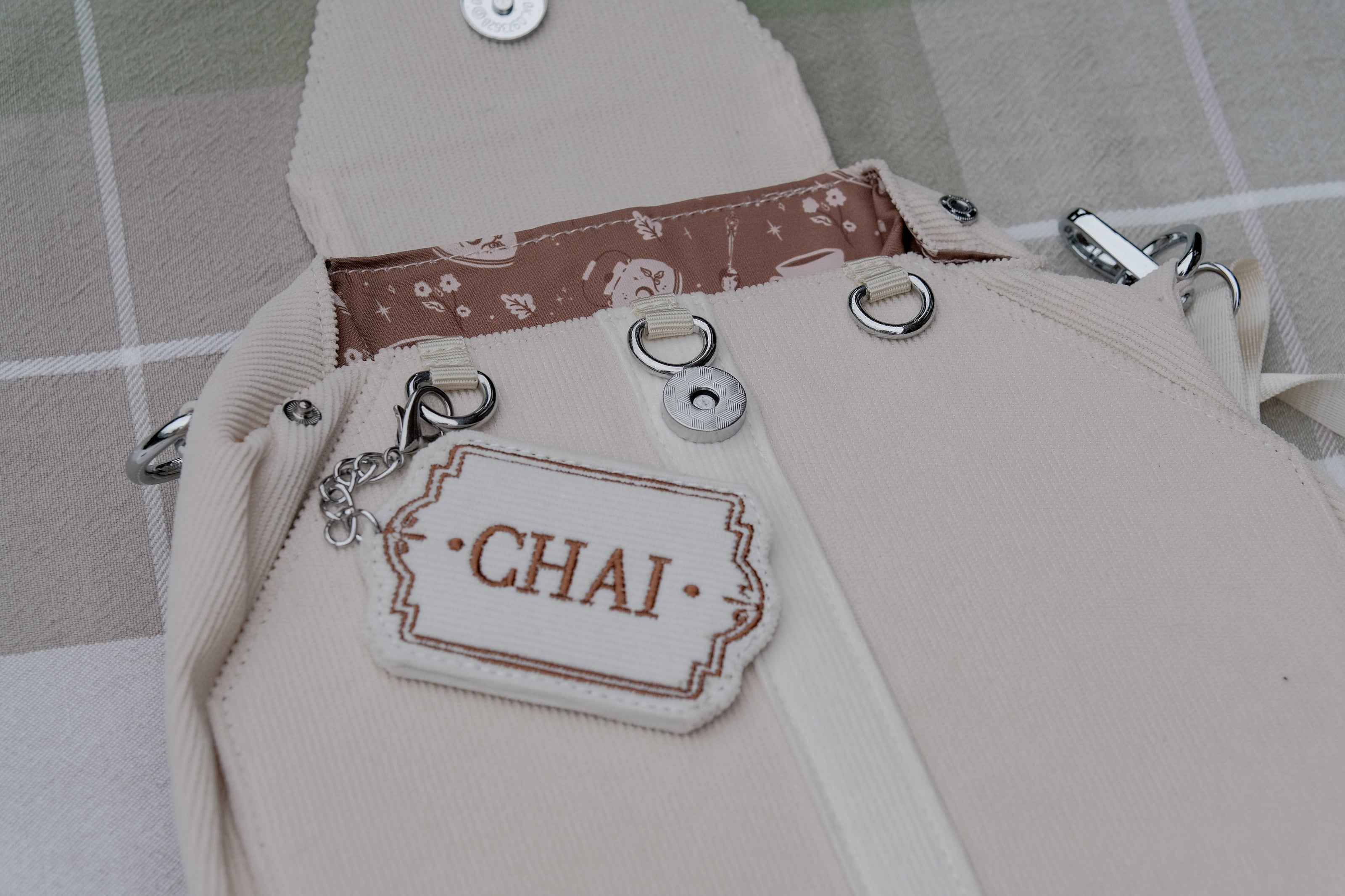 Ivory/cream and warm brown cotton corduroy purse, designed to resemble a chai tea bag, featuring the classic pyramid tea bag shape, adjustable matching strap, keychain rings, and a tag detail hanging from a strap. The bag features two inner pockets, one zipper and one padded, as well as 2 other unpadded pockets. Inner fabric is printed with a pattern of tea pots, bags, spoons, and cups, oak leaves, flowers, and stars on a soft brown background.