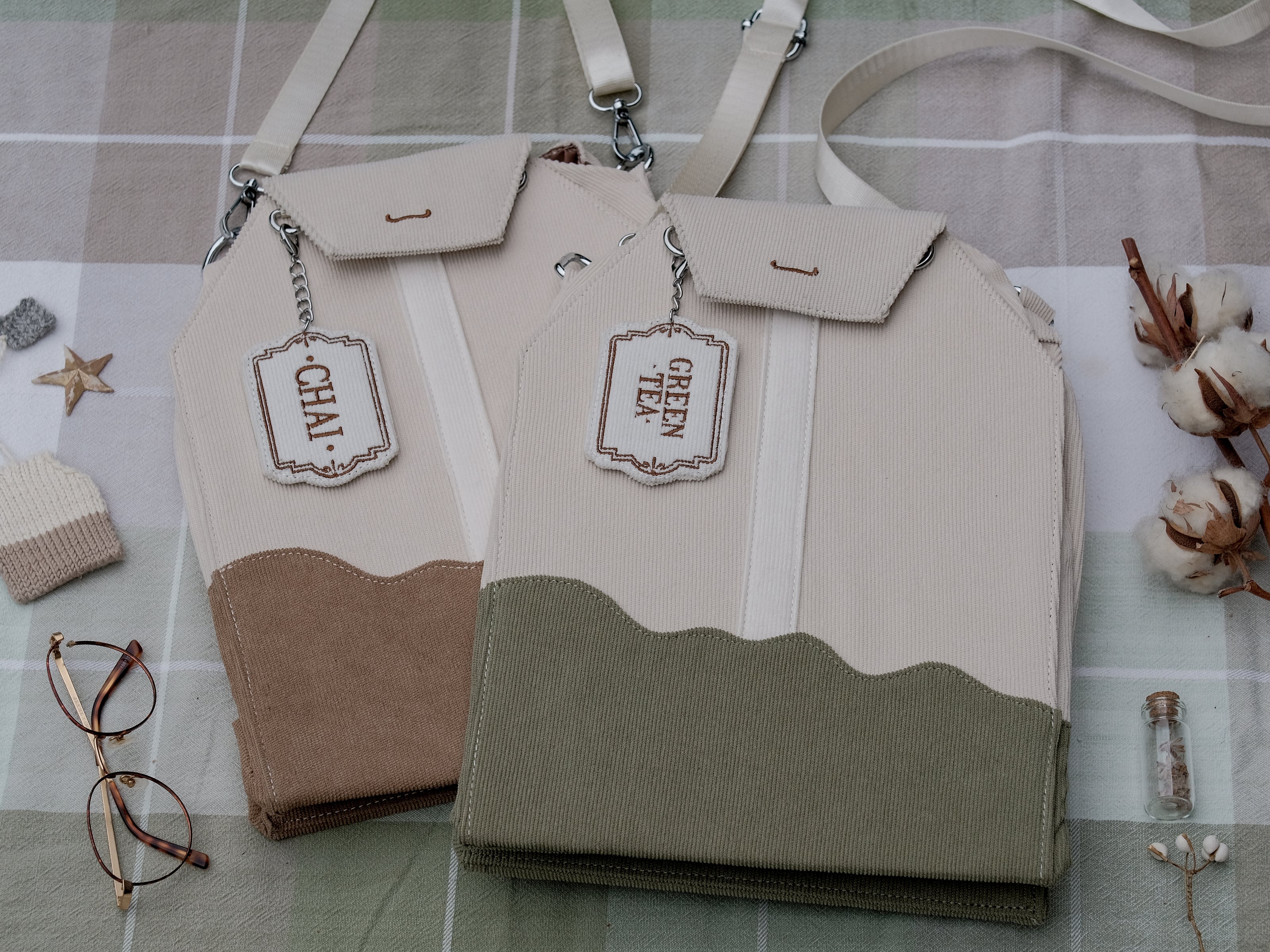 Ivory/cream and muted green cotton corduroy purse, designed to resemble a green tea bag, featuring the classic pyramid tea bag shape, adjustable matching strap, keychain rings, and a tag detail hanging from a strap. The bag features two inner pockets, one zipper and one padded, as well as 2 other unpadded pockets. Inner fabric is printed with a pattern of tea pots, bags, spoons, and mugs, bumble bees, flowers, and stars on a soft turquoise background.