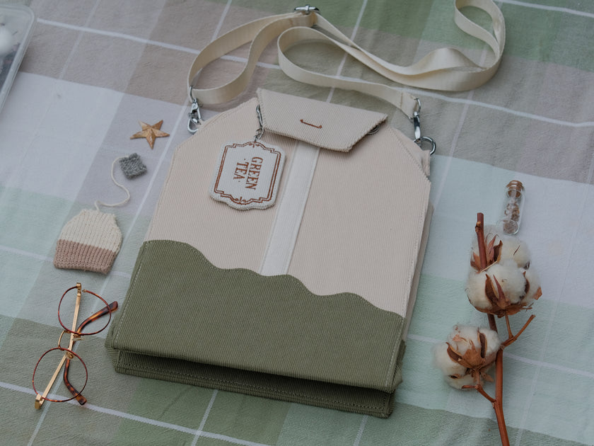 Ivory/cream and muted green cotton corduroy purse, designed to resemble a green tea bag, featuring the classic pyramid tea bag shape, adjustable matching strap, keychain rings, and a tag detail hanging from a strap. The bag features two inner pockets, one zipper and one padded, as well as 2 other unpadded pockets. Inner fabric is printed with a pattern of tea pots, bags, spoons, and mugs, bumble bees, flowers, and stars on a soft turquoise background.