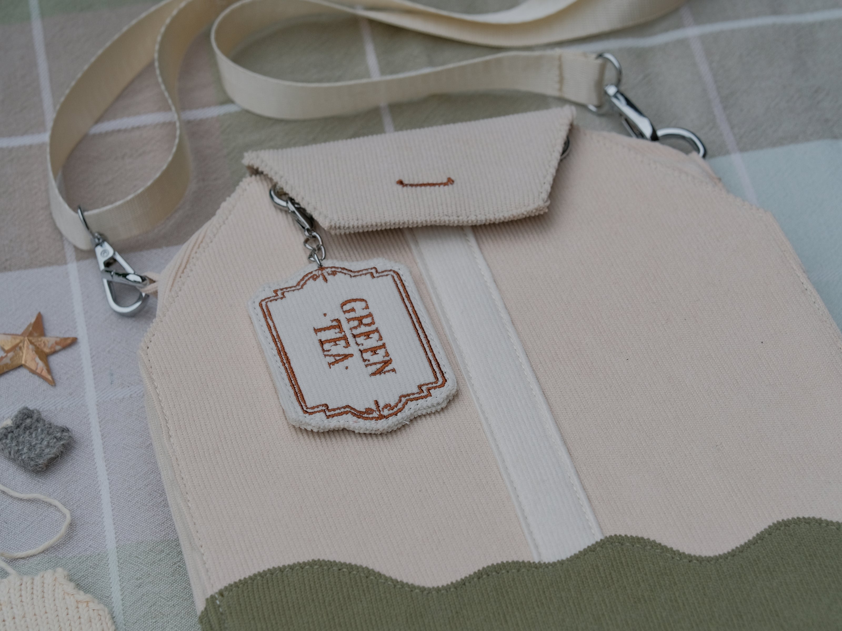 Ivory/cream and muted green cotton corduroy purse, designed to resemble a green tea bag, featuring the classic pyramid tea bag shape, adjustable matching strap, keychain rings, and a tag detail hanging from a strap. The bag features two inner pockets, one zipper and one padded, as well as 2 other unpadded pockets. Inner fabric is printed with a pattern of tea pots, bags, spoons, and mugs, bumble bees, flowers, and stars on a soft turquoise background.