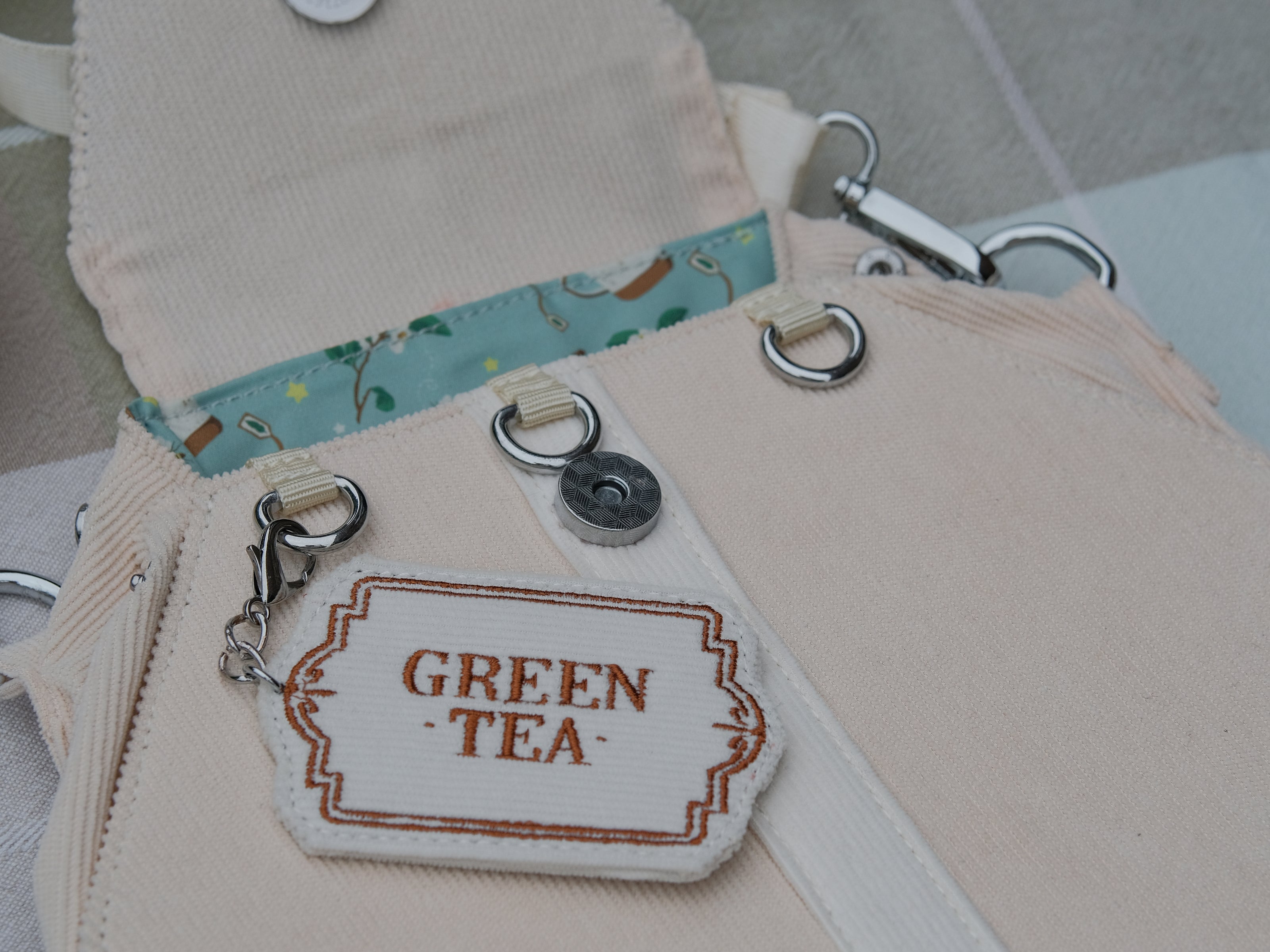 Ivory/cream and muted green cotton corduroy purse, designed to resemble a green tea bag, featuring the classic pyramid tea bag shape, adjustable matching strap, keychain rings, and a tag detail hanging from a strap. The bag features two inner pockets, one zipper and one padded, as well as 2 other unpadded pockets. Inner fabric is printed with a pattern of tea pots, bags, spoons, and mugs, bumble bees, flowers, and stars on a soft turquoise background.