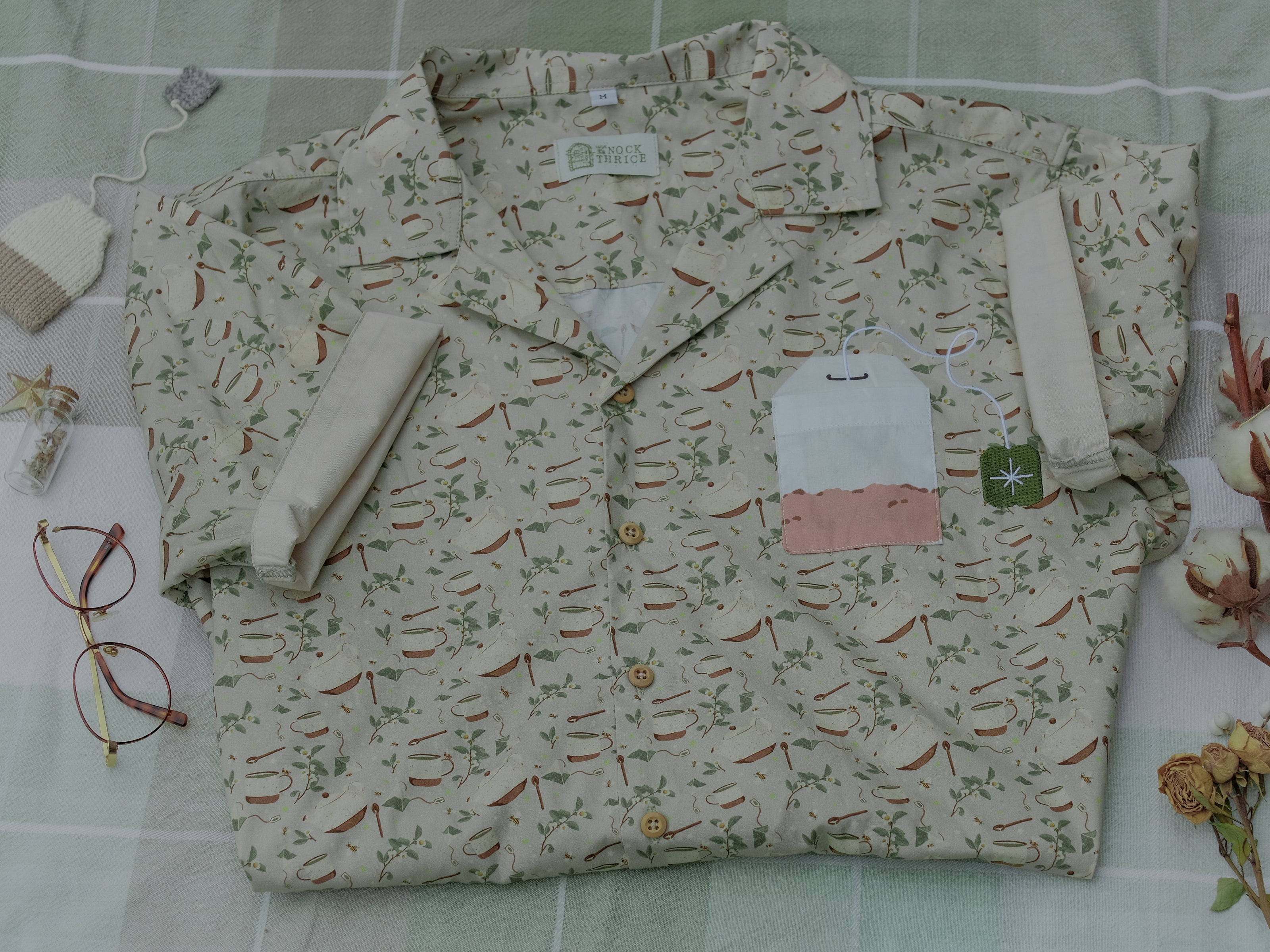A muted, warm green, short-sleeve button up t-shirt printed with a whimsical pattern of rustic tea cups and teapots, leafy sprigs, and buzzing bumble bees. The shirt has a pyramid tea bag pocket, as well as a relaxed fit and rolled cuffs.