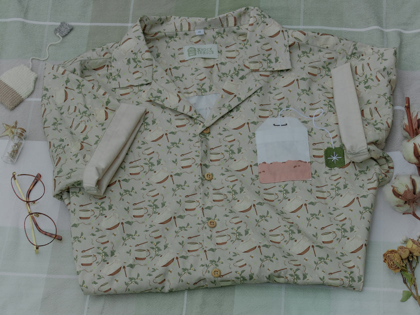 A muted, warm green, short-sleeve button up t-shirt printed with a whimsical pattern of rustic tea cups and teapots, leafy sprigs, and buzzing bumble bees. The shirt has a pyramid tea bag pocket, as well as a relaxed fit and rolled cuffs.