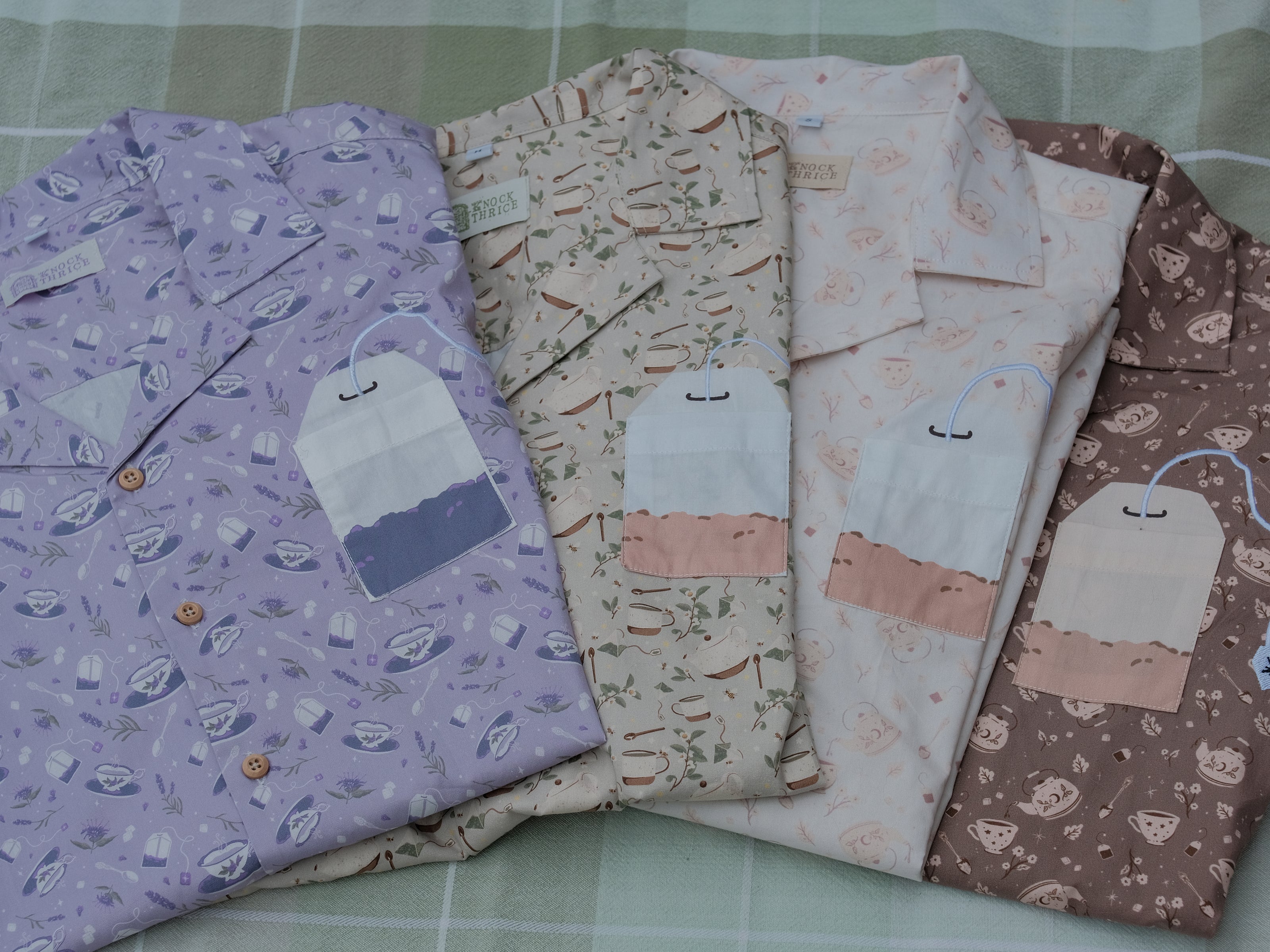 A muted, warm green, short-sleeve button up t-shirt printed with a whimsical pattern of rustic tea cups and teapots, leafy sprigs, and buzzing bumble bees. The shirt has a pyramid tea bag pocket, as well as a relaxed fit and rolled cuffs.