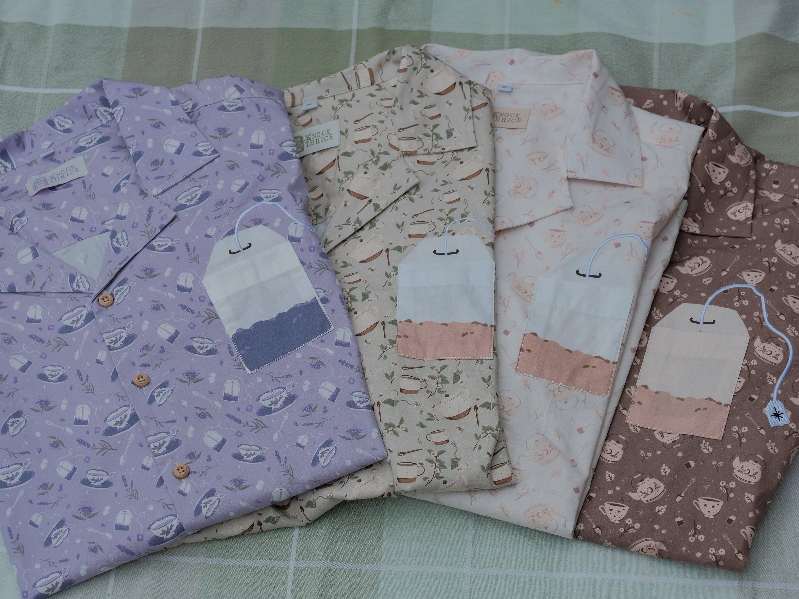 A muted purple, short-sleeve button up t-shirt printed with a whimsical pattern of fancy teacups, bergamot flowers, lavender sprigs, tea bags, spoons and sugar cubes. The shirt has a pyramid tea bag pocket, as well as a relaxed fit and rolled cuffs.