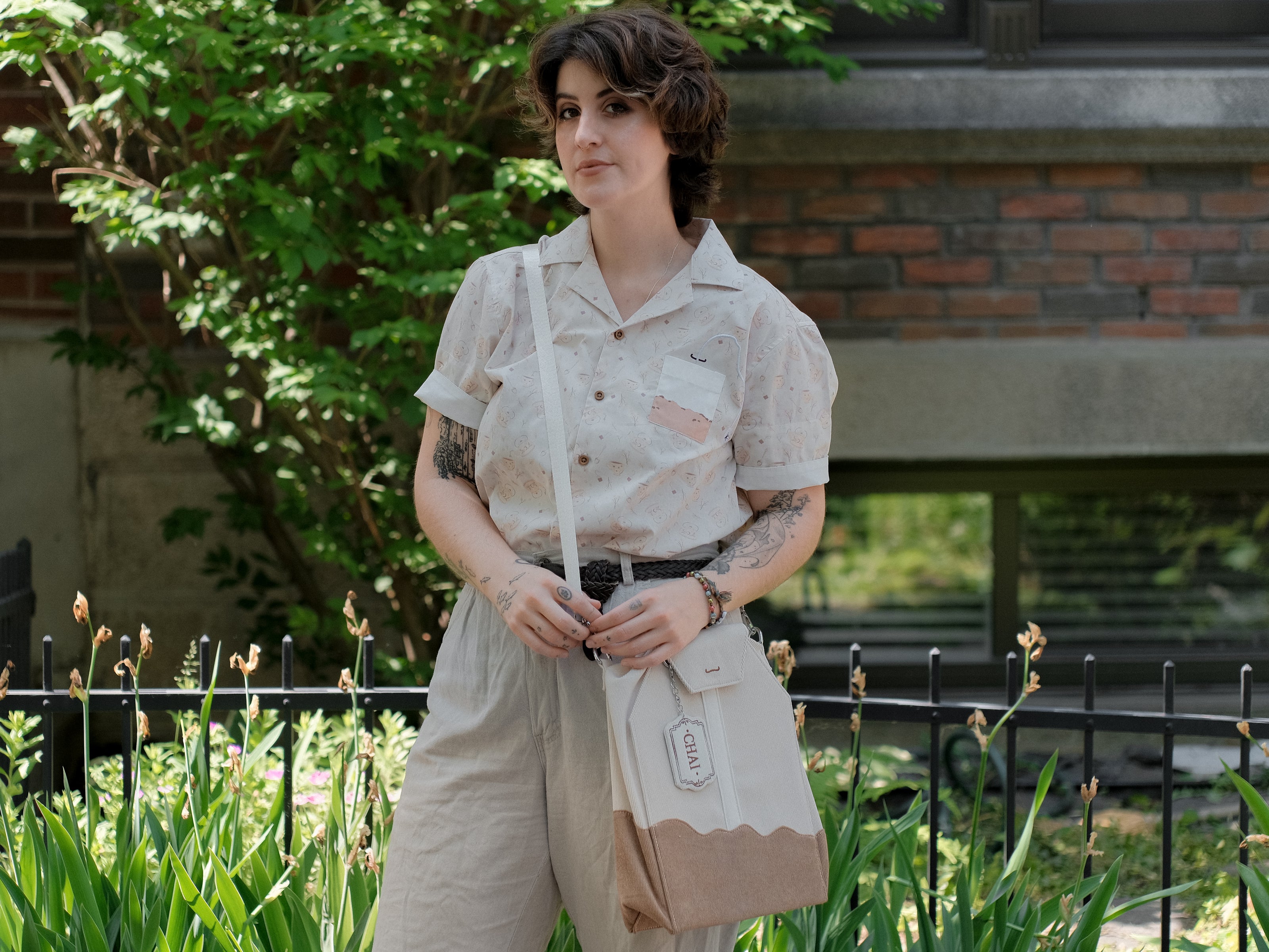 Ivory/cream and warm brown cotton corduroy purse, designed to resemble a chai tea bag, featuring the classic pyramid tea bag shape, adjustable matching strap, keychain rings, and a tag detail hanging from a strap. The bag features two inner pockets, one zipper and one padded, as well as 2 other unpadded pockets. Inner fabric is printed with a pattern of tea pots, bags, spoons, and cups, oak leaves, flowers, and stars on a soft brown background.