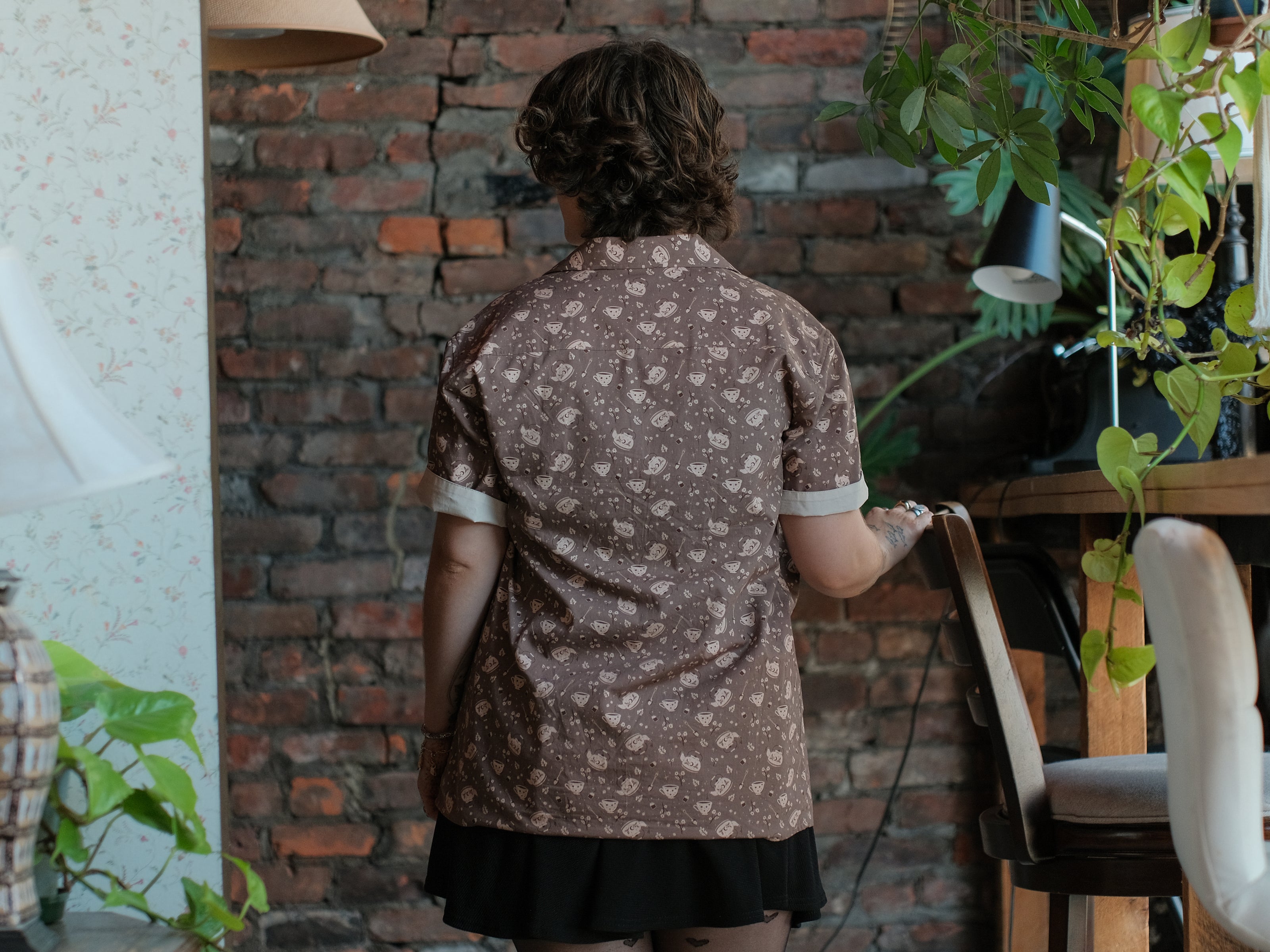 Warm, boba brown shade short-sleeve button up t-shirt printed with a whimsical pattern of the classic chubby teapot and matching cup, autumnal oak leaves, tea bag, spoon and sugar cubes. The shirt has a pyramid tea bag pocket, as well as a relaxed fit and rolled cuffs.