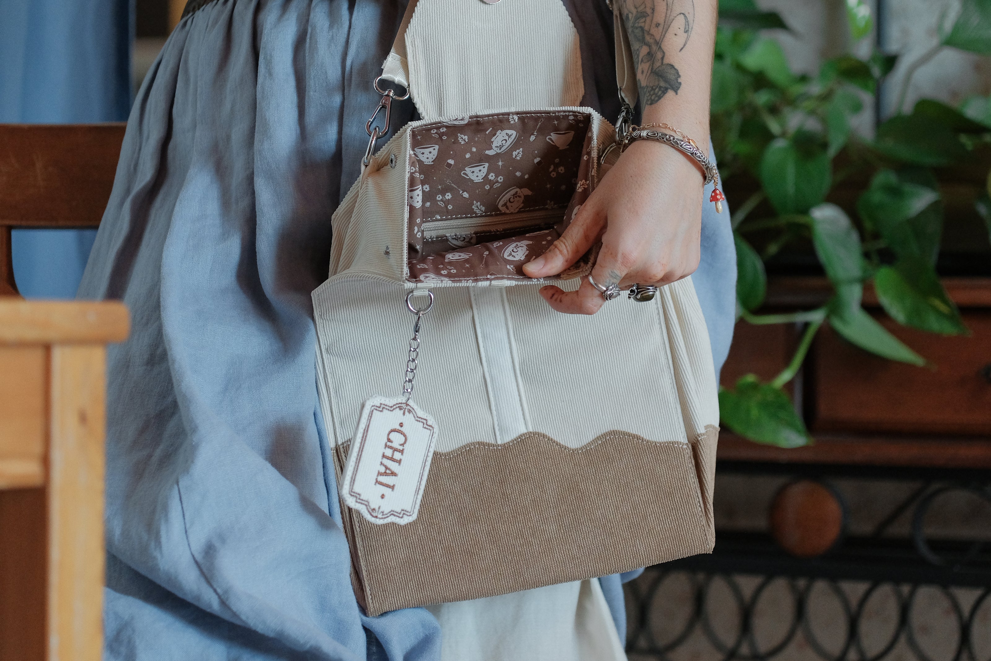Ivory/cream and warm brown cotton corduroy purse, designed to resemble a chai tea bag, featuring the classic pyramid tea bag shape, adjustable matching strap, keychain rings, and a tag detail hanging from a strap. The bag features two inner pockets, one zipper and one padded, as well as 2 other unpadded pockets. Inner fabric is printed with a pattern of tea pots, bags, spoons, and cups, oak leaves, flowers, and stars on a soft brown background.