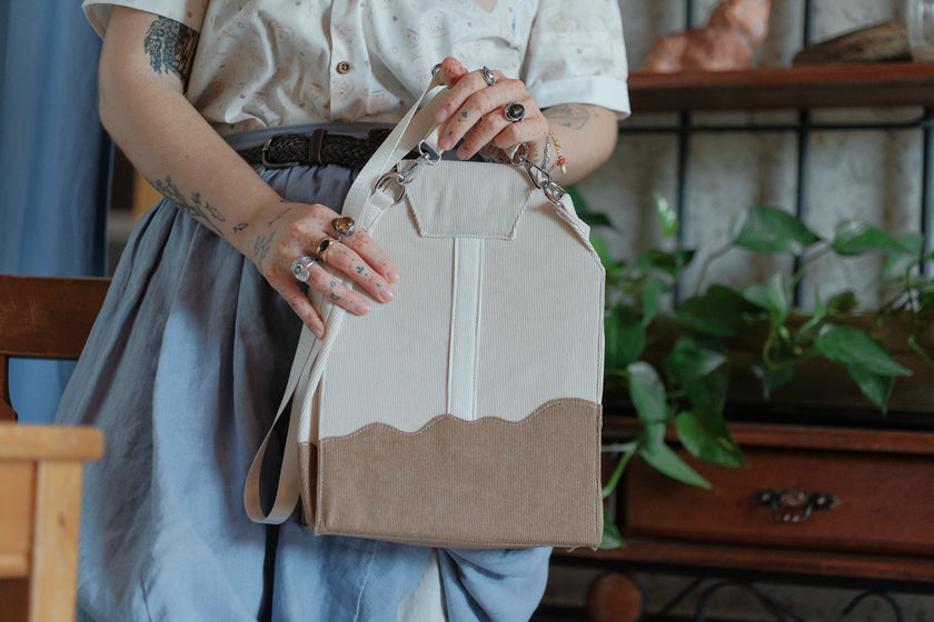 IIvory/cream and warm brown cotton corduroy purse, designed to resemble a chai tea bag, featuring the classic pyramid tea bag shape, adjustable matching strap, keychain rings, and a tag detail hanging from a strap. The bag features two inner pockets, one zipper and one padded, as well as 2 other unpadded pockets. Inner fabric is printed with a pattern of tea pots, bags, spoons, and cups, oak leaves, flowers, and stars on a soft brown background.
