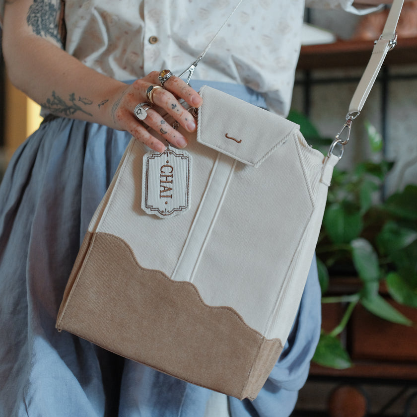 Ivory/cream and warm brown cotton corduroy purse, designed to resemble a chai tea bag, featuring the classic pyramid tea bag shape, adjustable matching strap, keychain rings, and a tag detail hanging from a strap. The bag features two inner pockets, one zipper and one padded, as well as 2 other unpadded pockets. Inner fabric is printed with a pattern of tea pots, bags, spoons, and cups, oak leaves, flowers, and stars on a soft brown background.