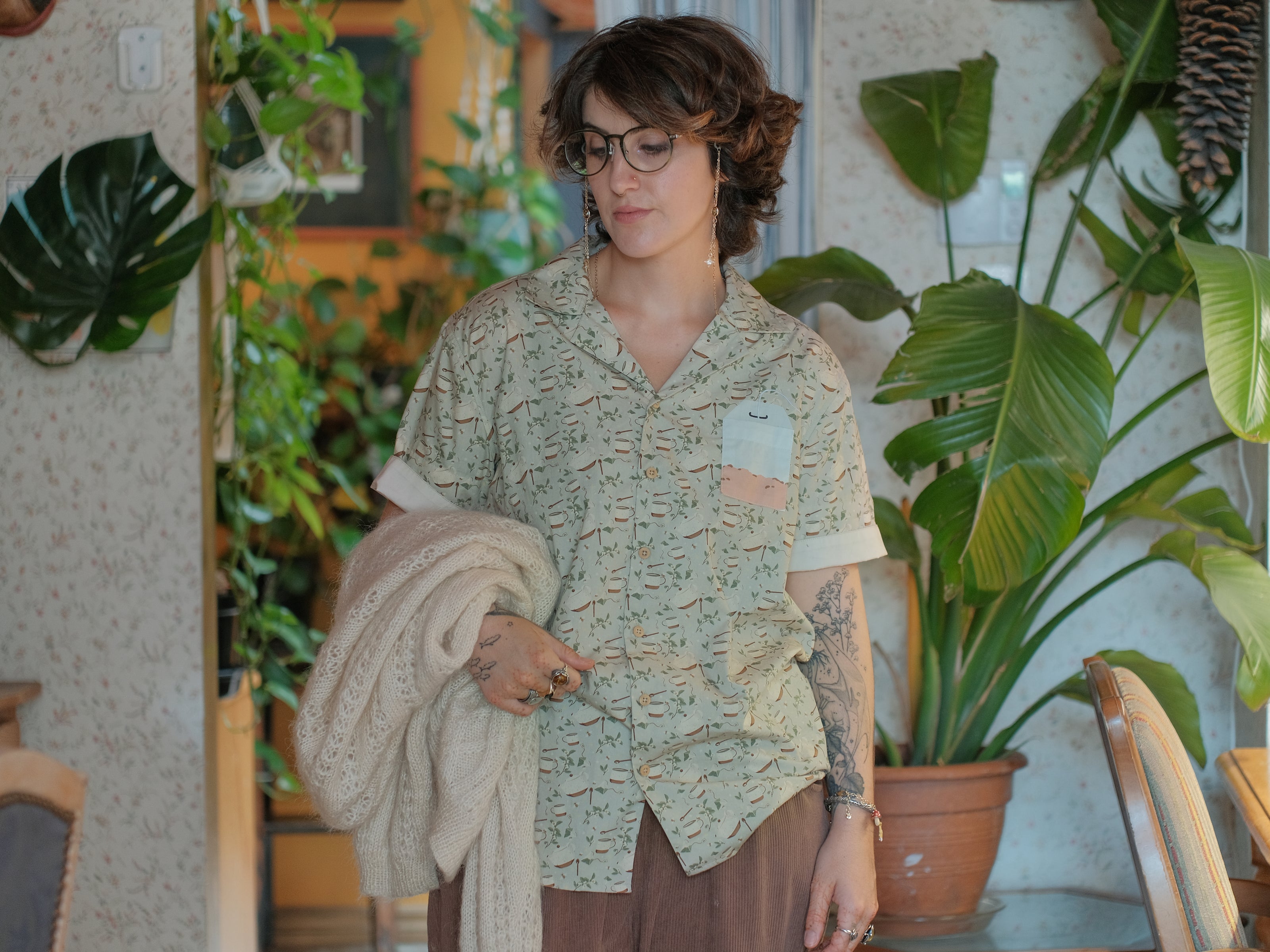 A muted, warm green, short-sleeve button up t-shirt printed with a whimsical pattern of rustic tea cups and teapots, leafy sprigs, and buzzing bumble bees. The shirt has a pyramid tea bag pocket, as well as a relaxed fit and rolled cuffs.
