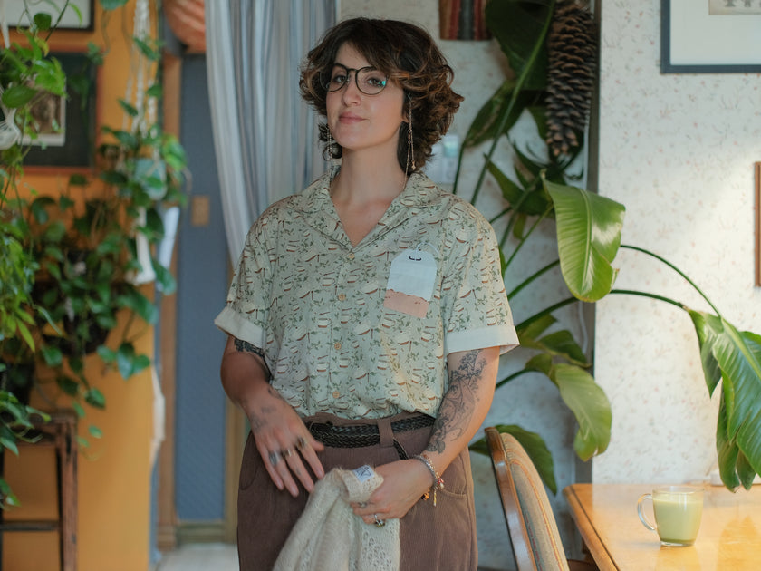 A muted, warm green, short-sleeve button up t-shirt printed with a whimsical pattern of rustic tea cups and teapots, leafy sprigs, and buzzing bumble bees. The shirt has a pyramid tea bag pocket, as well as a relaxed fit and rolled cuffs.