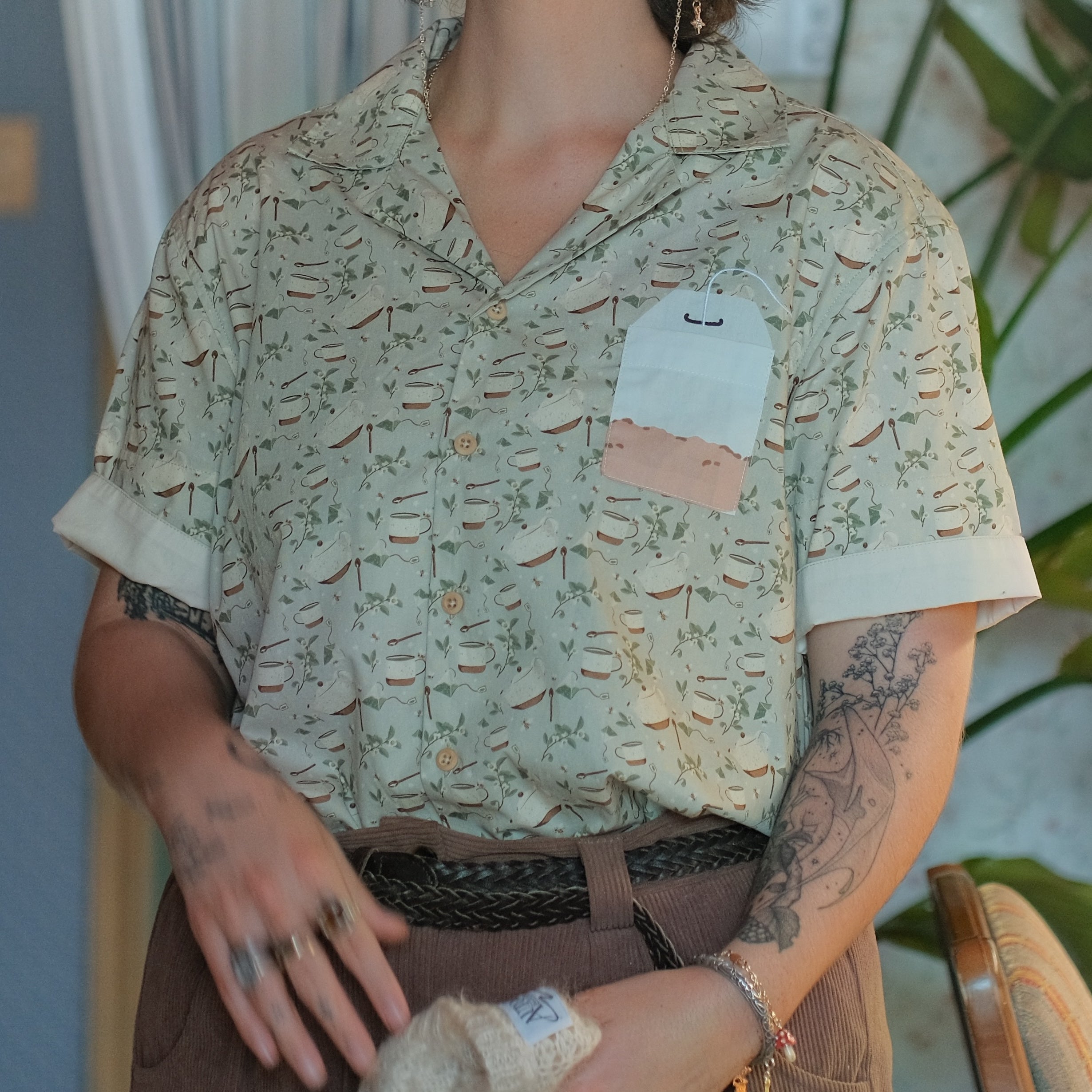 A muted, warm green, short-sleeve button up t-shirt printed with a whimsical pattern of rustic tea cups and teapots, leafy sprigs, and buzzing bumble bees. The shirt has a pyramid tea bag pocket, as well as a relaxed fit and rolled cuffs.