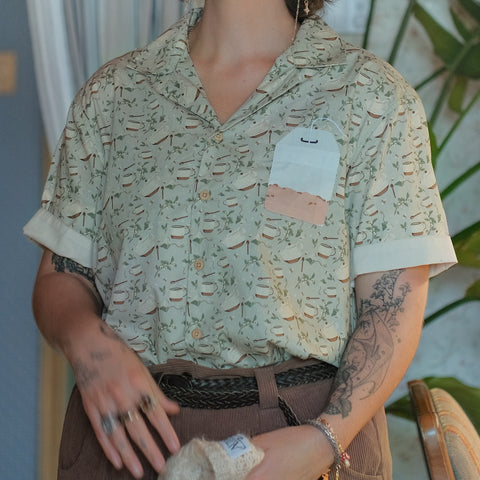 A muted, warm green, short-sleeve button up t-shirt printed with a whimsical pattern of rustic tea cups and teapots, leafy sprigs, and buzzing bumble bees. The shirt has a pyramid tea bag pocket, as well as a relaxed fit and rolled cuffs.
