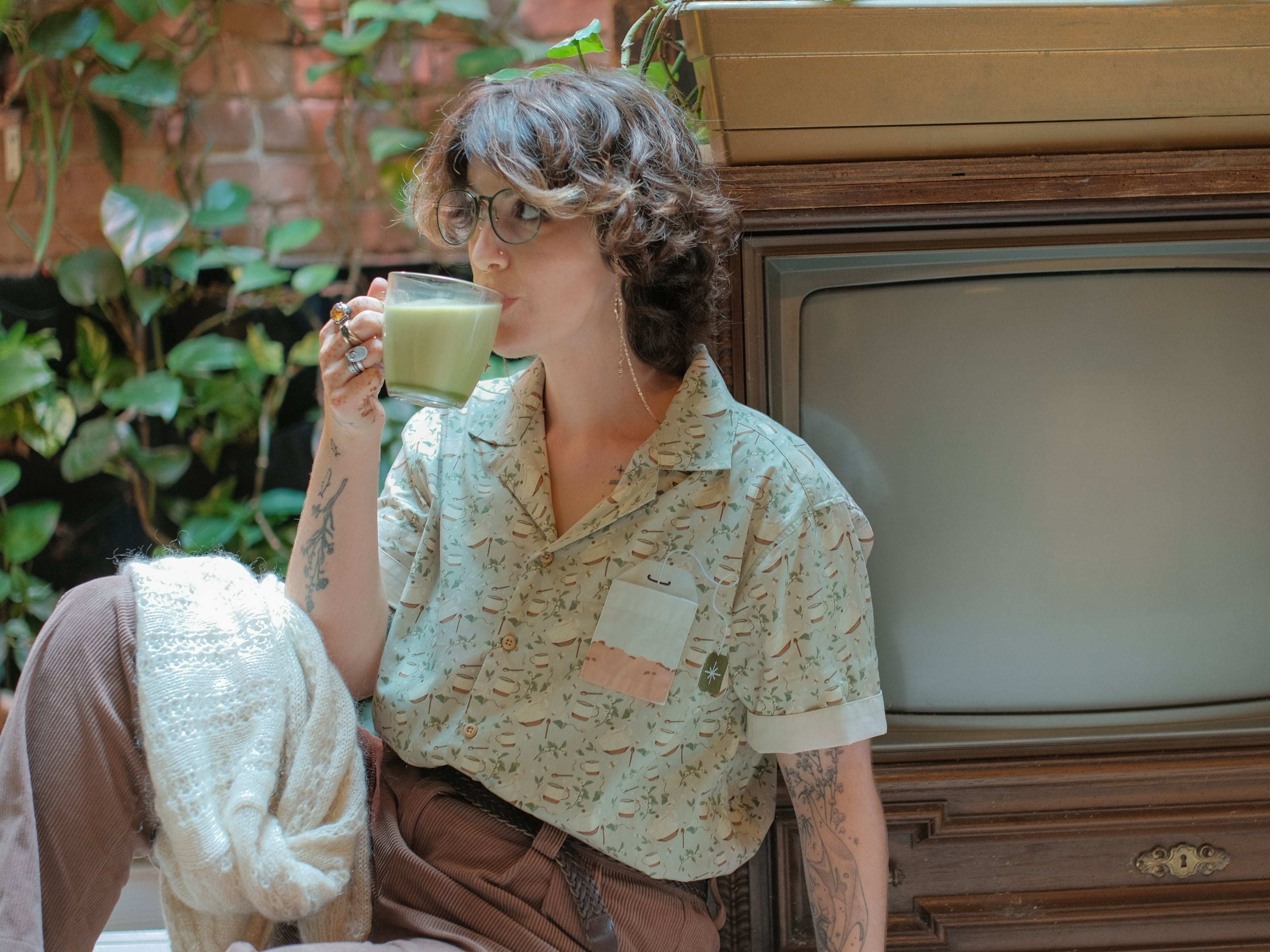 A muted, warm green, short-sleeve button up t-shirt printed with a whimsical pattern of rustic tea cups and teapots, leafy sprigs, and buzzing bumble bees. The shirt has a pyramid tea bag pocket, as well as a relaxed fit and rolled cuffs.