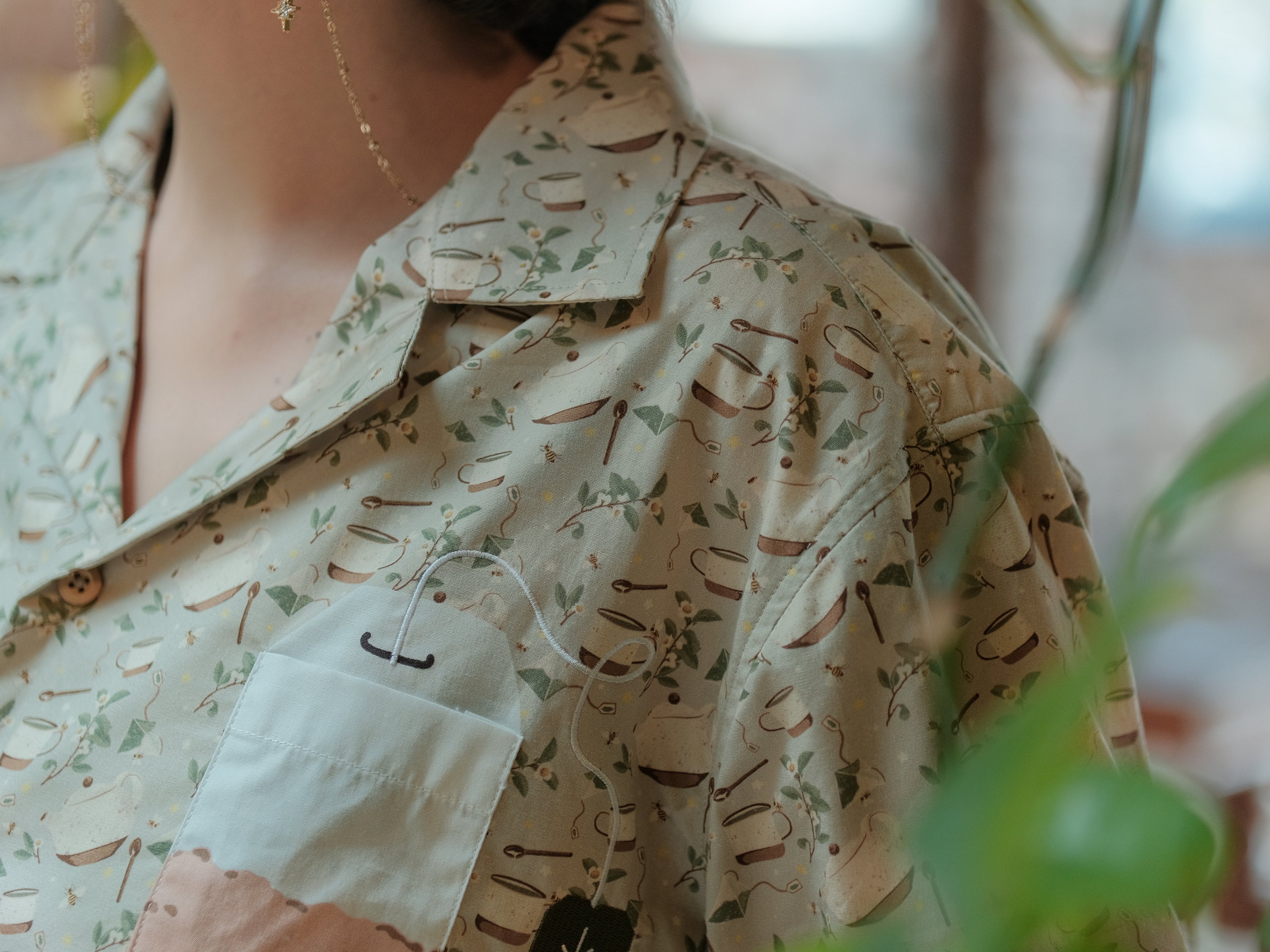 A muted, warm green, short-sleeve button up t-shirt printed with a whimsical pattern of rustic tea cups and teapots, leafy sprigs, and buzzing bumble bees. The shirt has a pyramid tea bag pocket, as well as a relaxed fit and rolled cuffs.