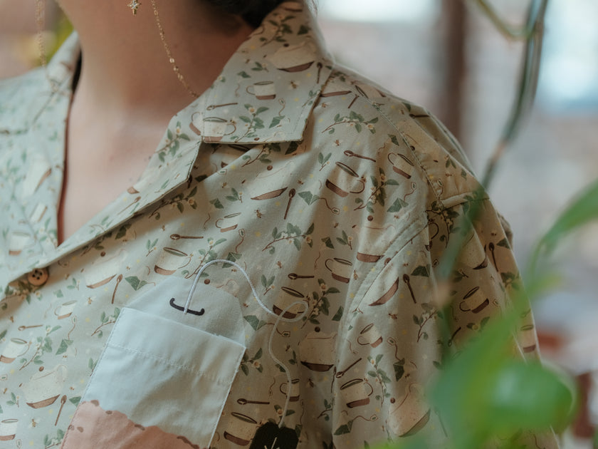 A muted, warm green, short-sleeve button up t-shirt printed with a whimsical pattern of rustic tea cups and teapots, leafy sprigs, and buzzing bumble bees. The shirt has a pyramid tea bag pocket, as well as a relaxed fit and rolled cuffs.
