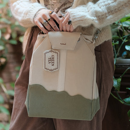 Ivory/cream and muted green cotton corduroy purse, designed to resemble a green tea bag, featuring the classic pyramid tea bag shape, adjustable matching strap, keychain rings, and a tag detail hanging from a strap. The bag features two inner pockets, one zipper and one padded, as well as 2 other unpadded pockets. Inner fabric is printed with a pattern of tea pots, bags, spoons, and mugs, bumble bees, flowers, and stars on a soft turquoise background.