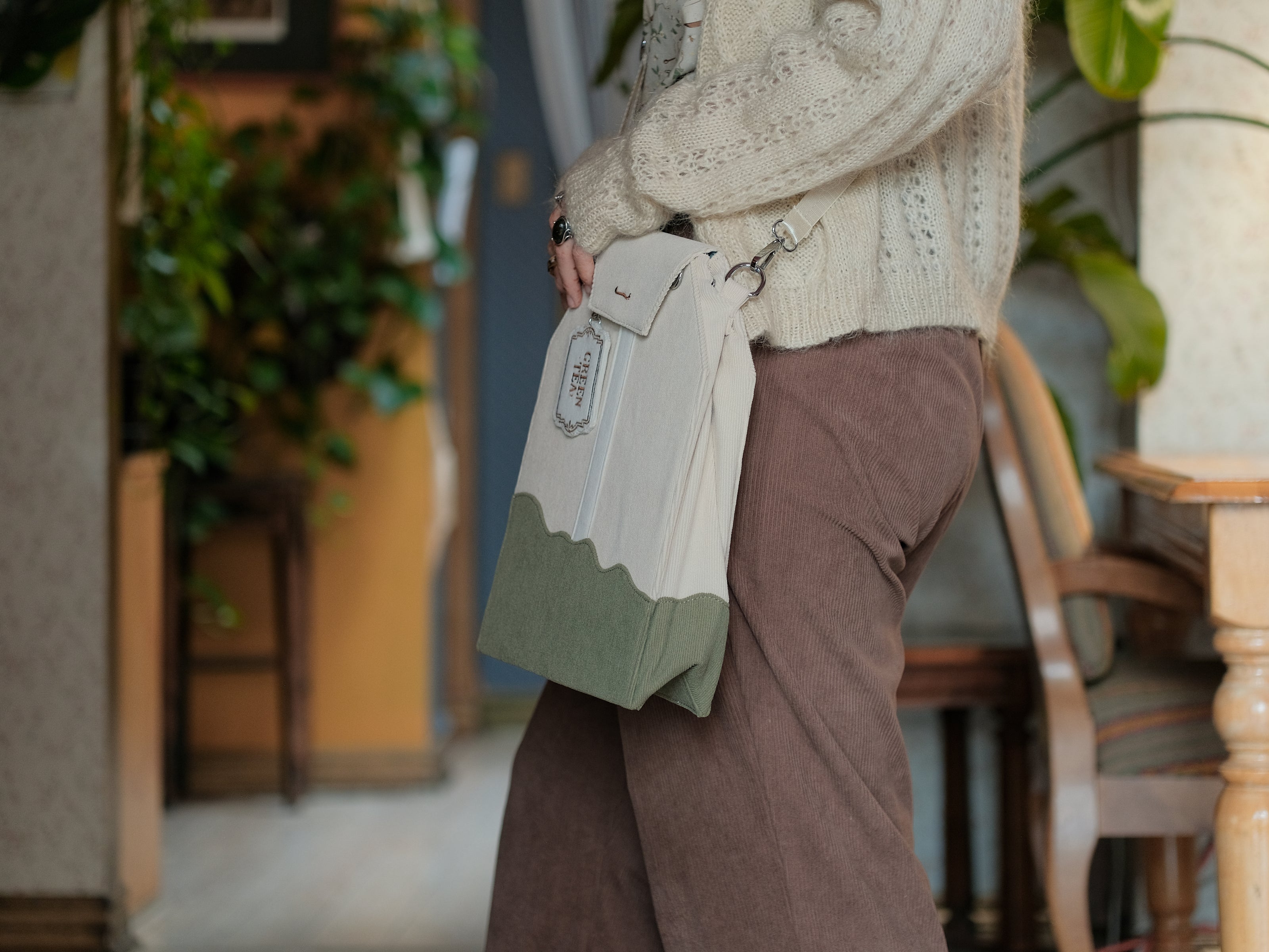 Ivory/cream and muted green cotton corduroy purse, designed to resemble a green tea bag, featuring the classic pyramid tea bag shape, adjustable matching strap, keychain rings, and a tag detail hanging from a strap. The bag features two inner pockets, one zipper and one padded, as well as 2 other unpadded pockets. Inner fabric is printed with a pattern of tea pots, bags, spoons, and mugs, bumble bees, flowers, and stars on a soft turquoise background.