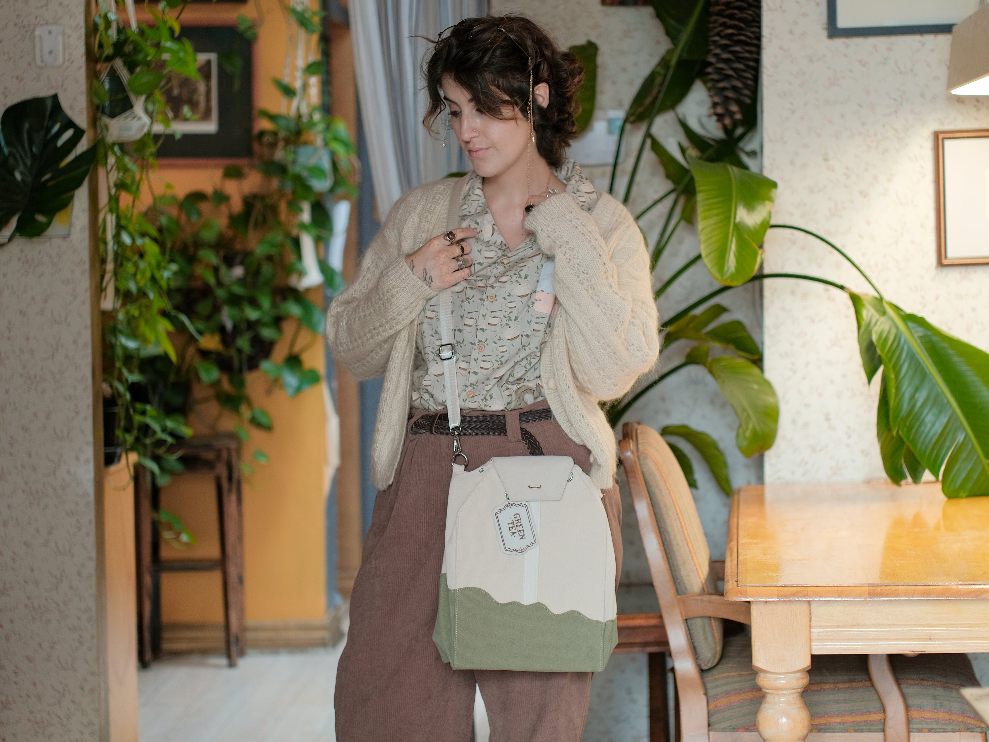 Ivory/cream and muted green cotton corduroy purse, designed to resemble a green tea bag, featuring the classic pyramid tea bag shape, adjustable matching strap, keychain rings, and a tag detail hanging from a strap. The bag features two inner pockets, one zipper and one padded, as well as 2 other unpadded pockets. Inner fabric is printed with a pattern of tea pots, bags, spoons, and mugs, bumble bees, flowers, and stars on a soft turquoise background.