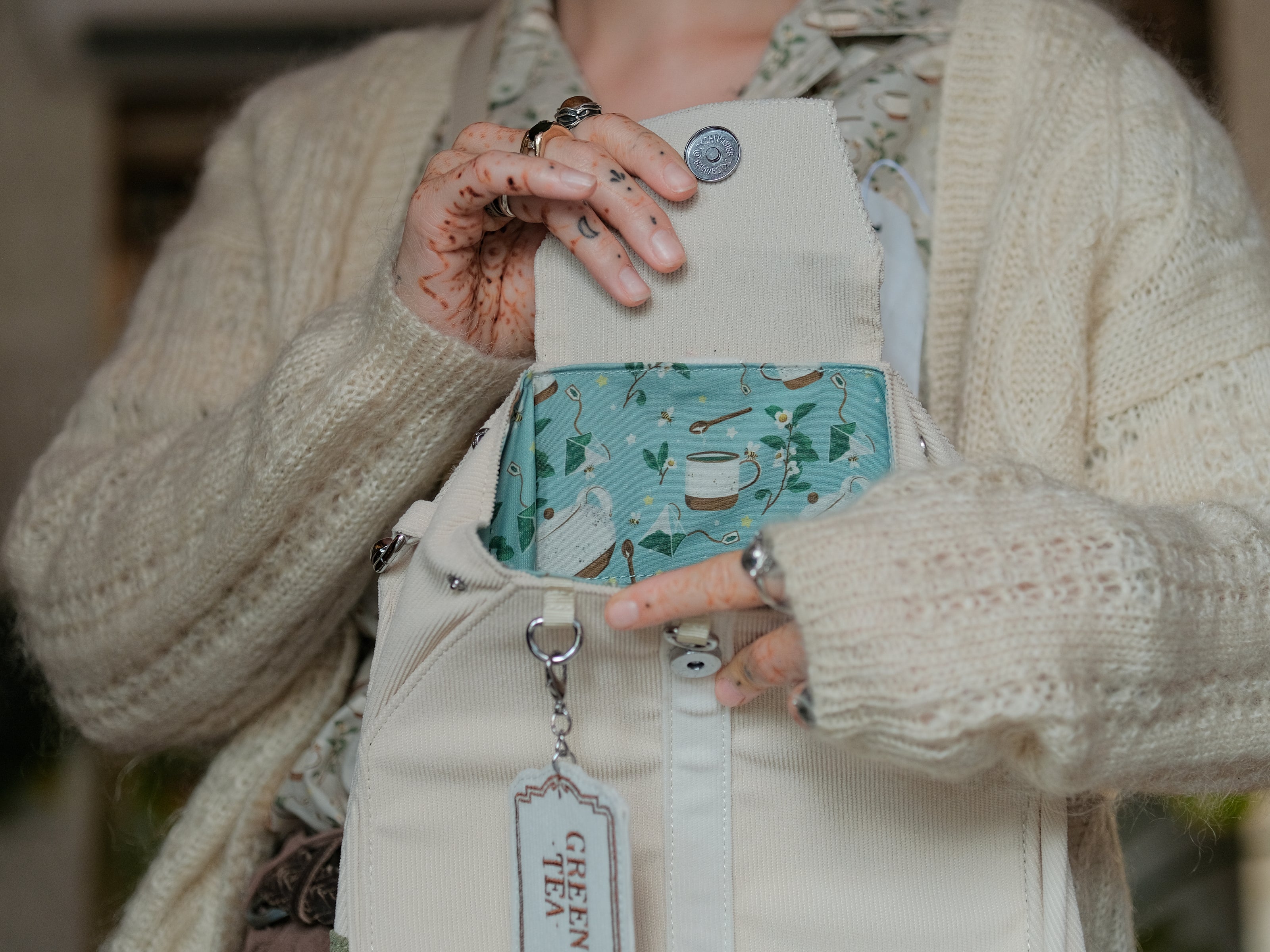 Ivory/cream and muted green cotton corduroy purse, designed to resemble a green tea bag, featuring the classic pyramid tea bag shape, adjustable matching strap, keychain rings, and a tag detail hanging from a strap. The bag features two inner pockets, one zipper and one padded, as well as 2 other unpadded pockets. Inner fabric is printed with a pattern of tea pots, bags, spoons, and mugs, bumble bees, flowers, and stars on a soft turquoise background.