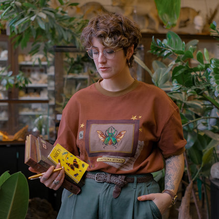 A rusty, vintage orange cotton T-shirt featuring an embroidered stag-horn beetle design framed with vintage-style details, handwritten notes, celestial accents, and latin nomenclature.